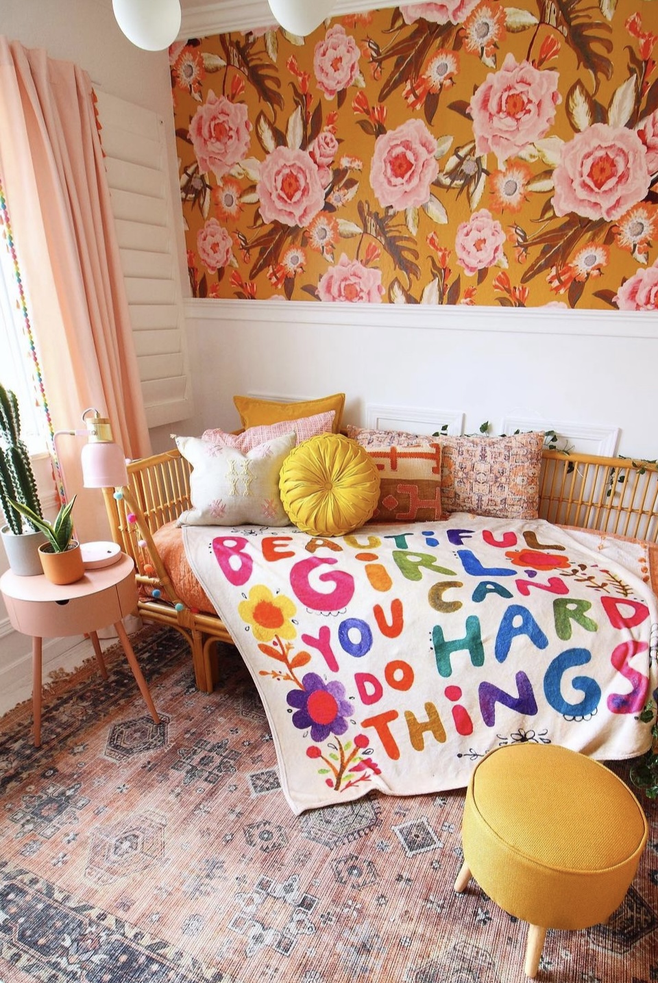 Bedroom with floral wallpaper, pink curtains, colorful pillows, and a blanket with colorful text, yellow ottoman, and potted plant.