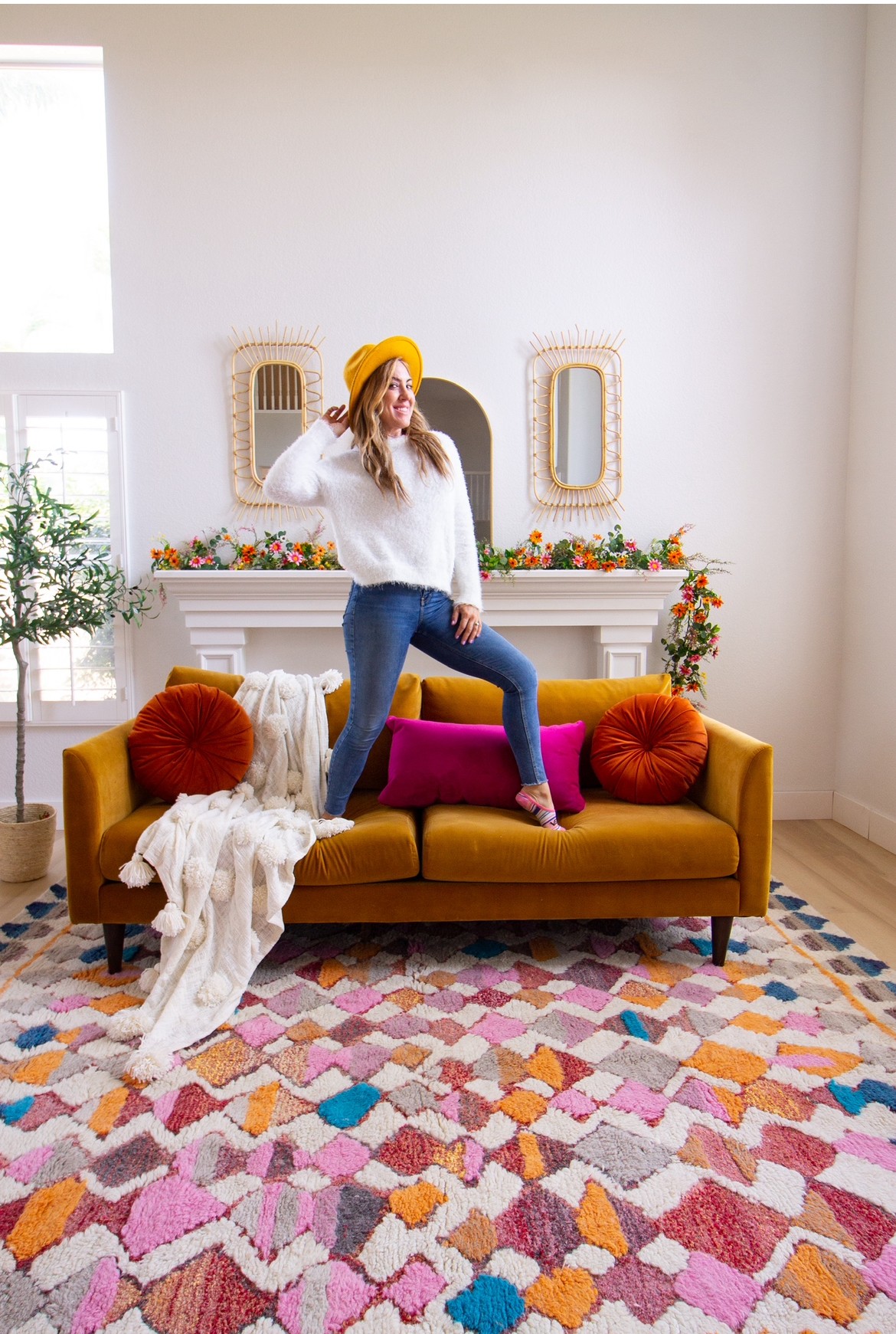 Woman in white sweater, yellow hat, and pink boots standing on a yellow sofa with red pillows in a bright room.