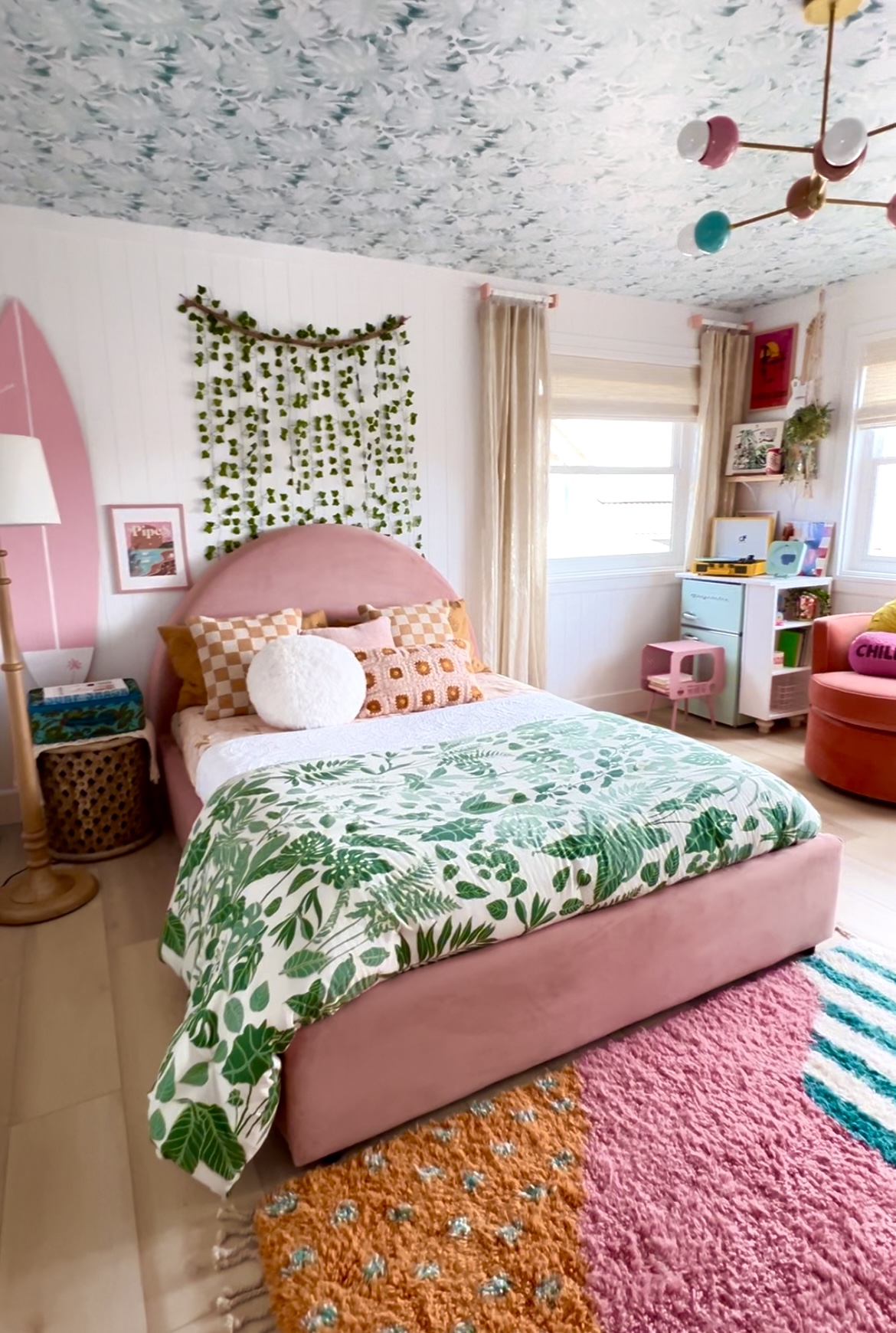 Bedroom with pink bed, green leafy bedding, colorful rug, and window with curtains, decorated with plants and artwork.
