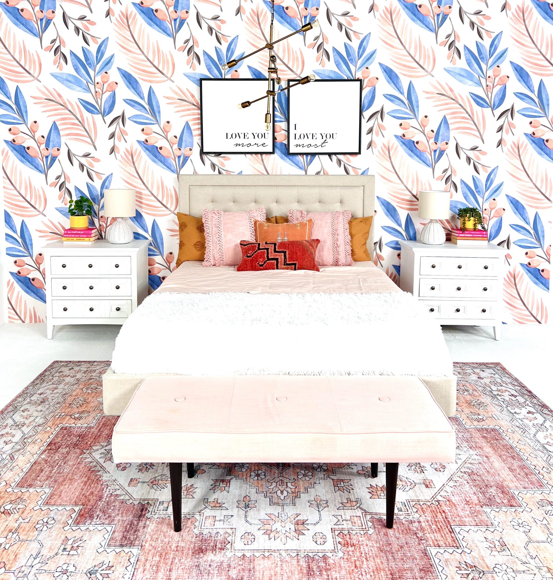 Bedroom with floral wallpaper, bed with pink and orange pillows, two white nightstands with plants, and a pink bench.