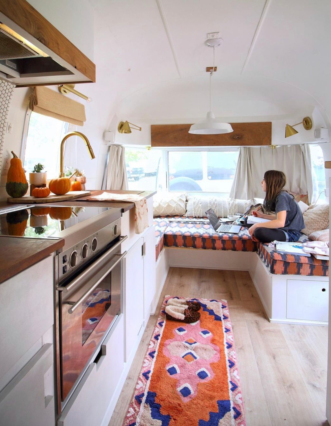Person sitting on a window seat in a cozy kitchen with a colorful rug, white cabinets, and a window with curtains.