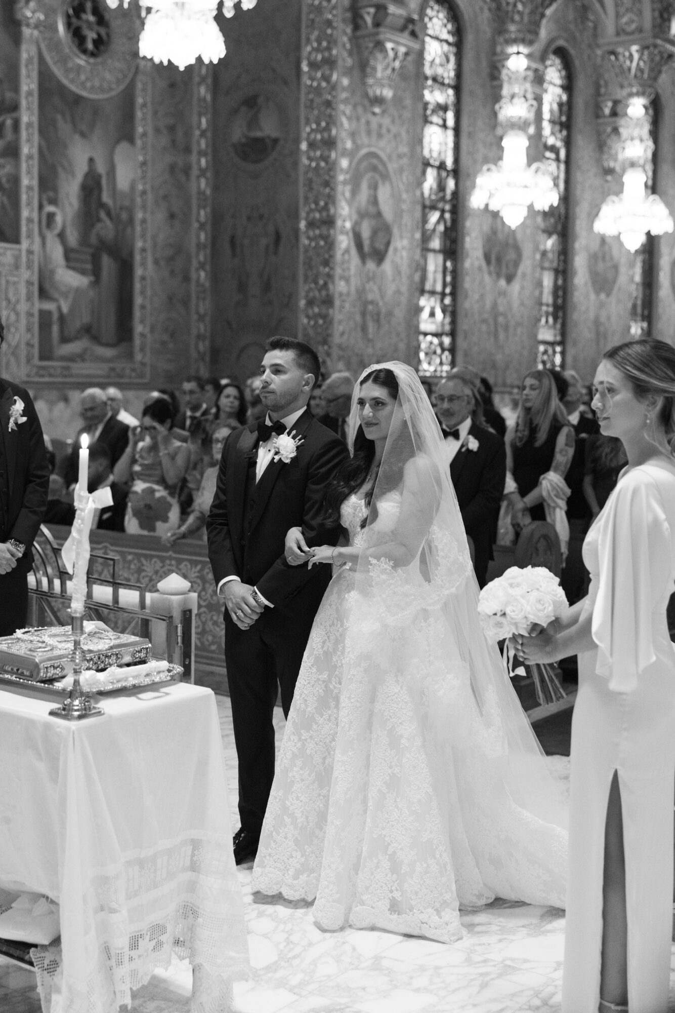 Bride and groom standing together during wedding ceremony inside ornate church, surrounded by guests, with a table and cake nearby.