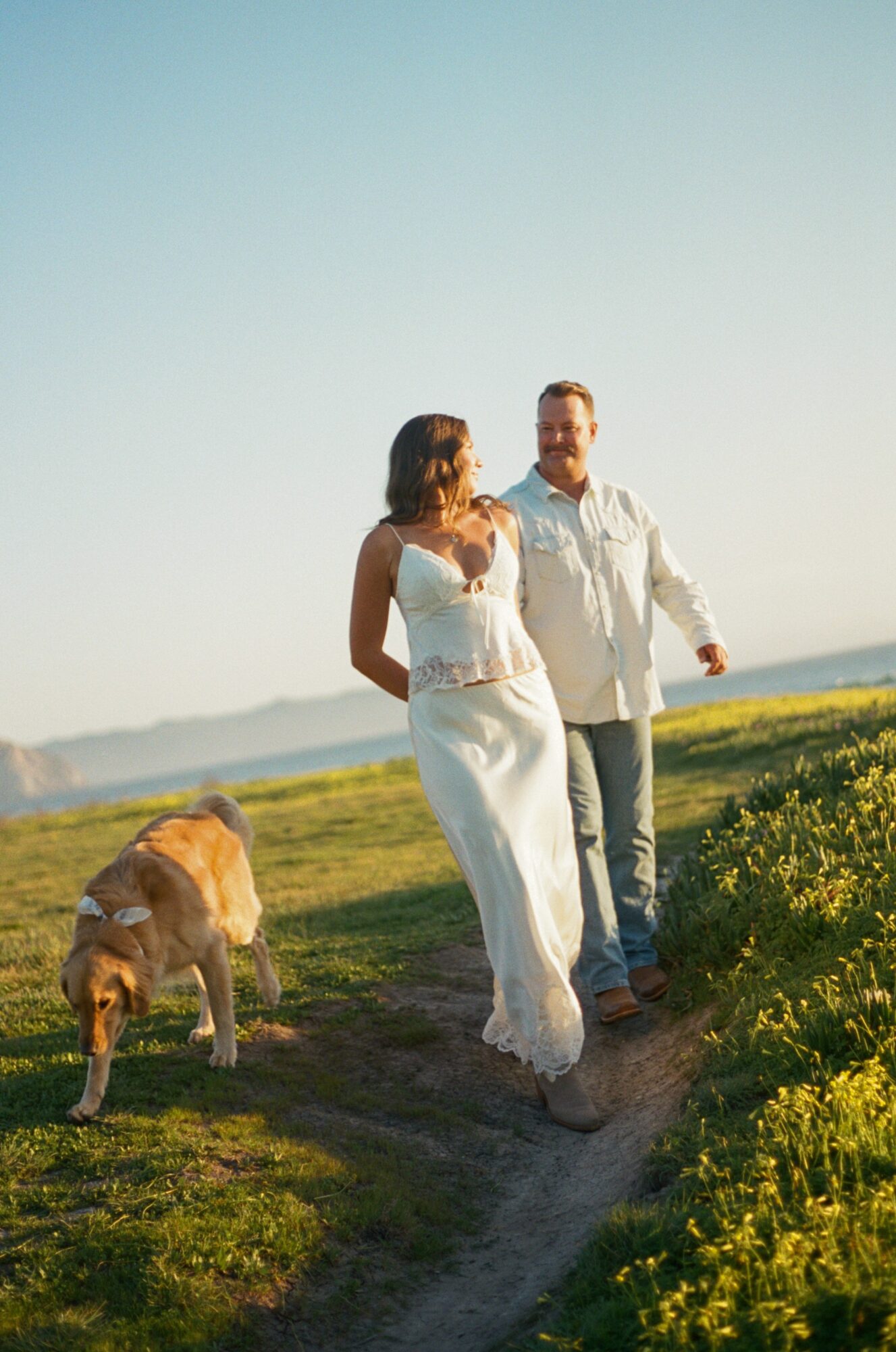 A couple walking outdoors with a dog, grassy area, and clear sky in the background.