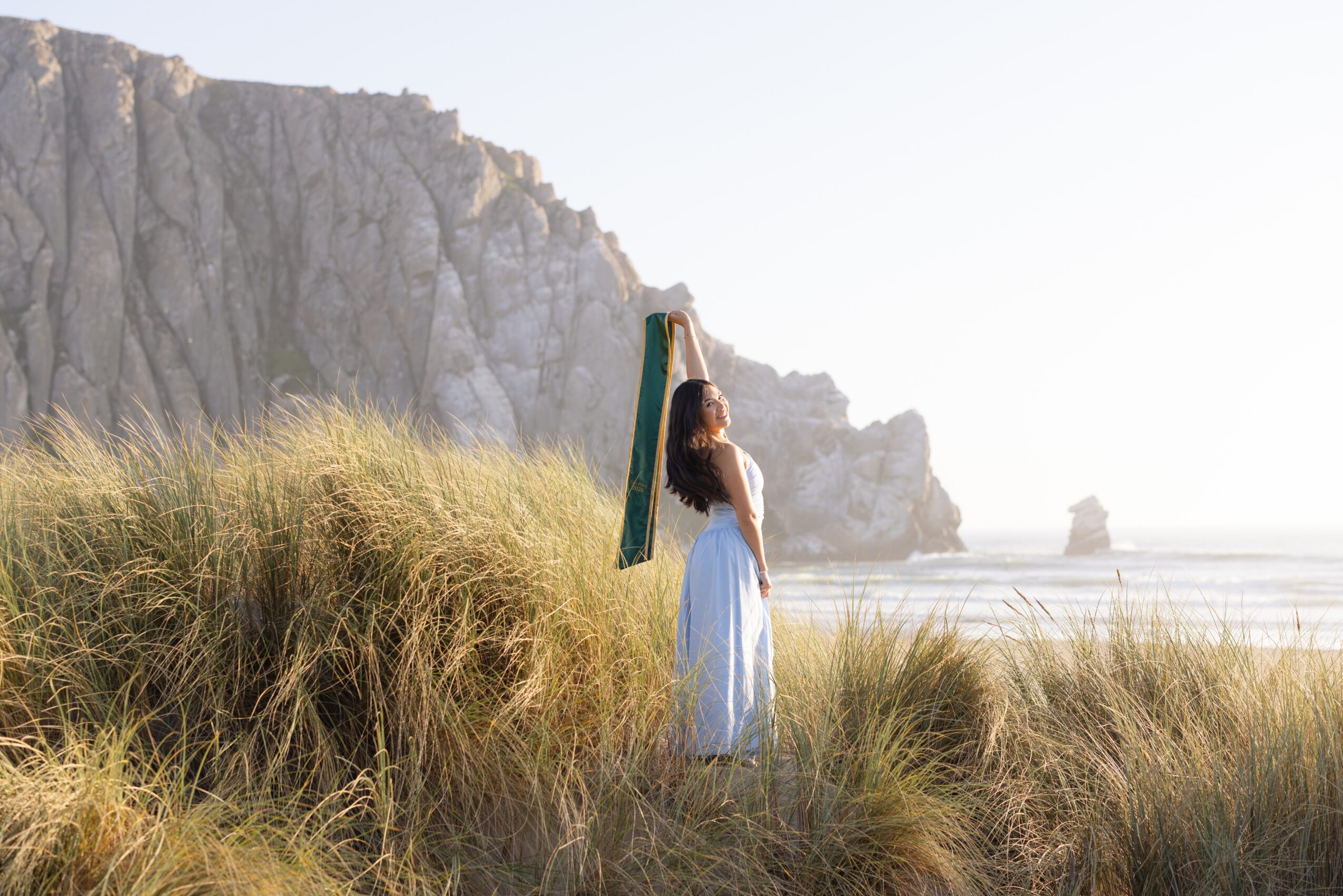 Woman in a white dress standing on grassy dunes near cliffs and the ocean, holding a green and yellow umbrella, facing away.