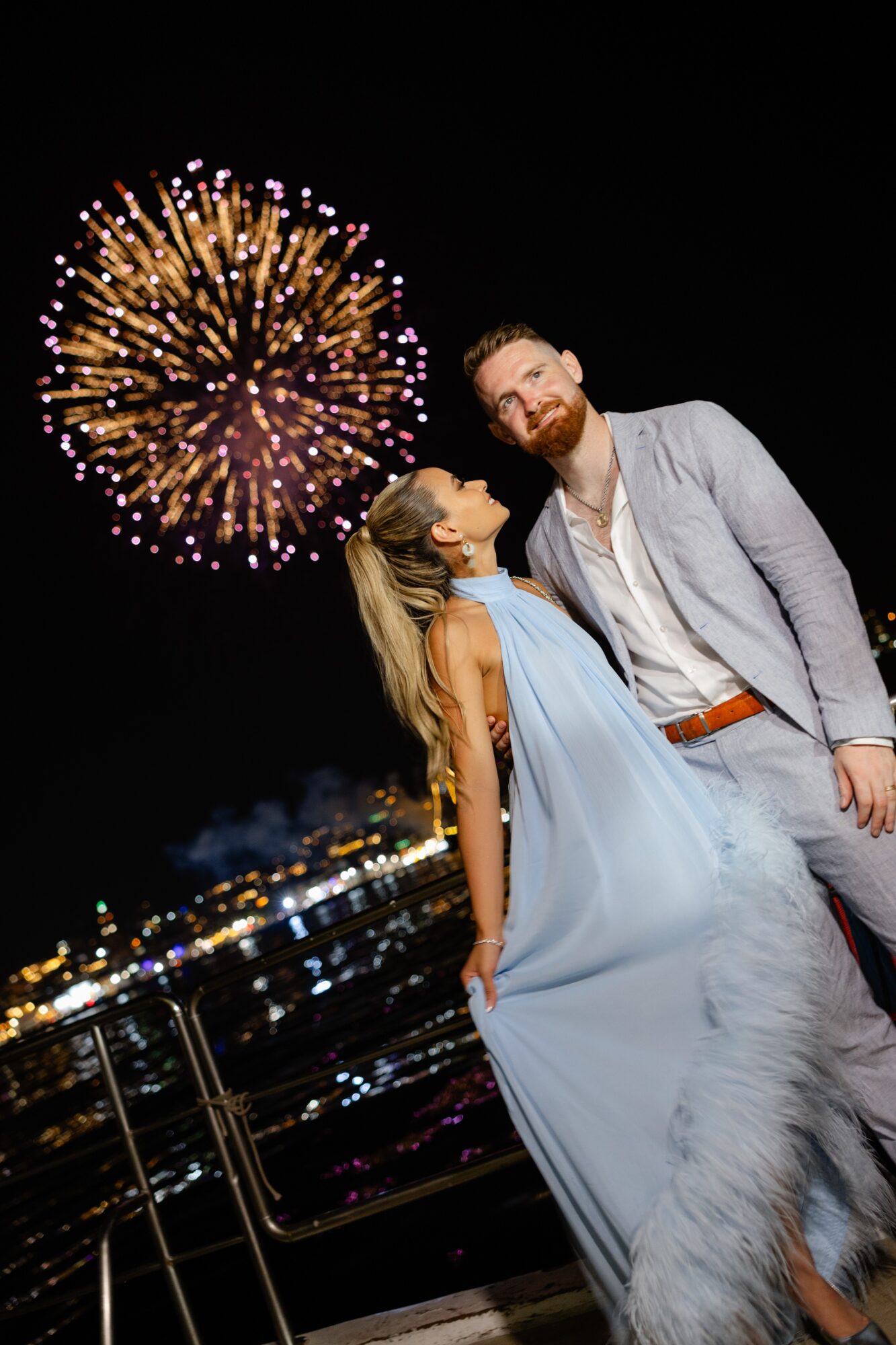 A couple stands near a railing at night with fireworks in the sky behind them, city lights below, woman in a light blue dress, man in a suit.