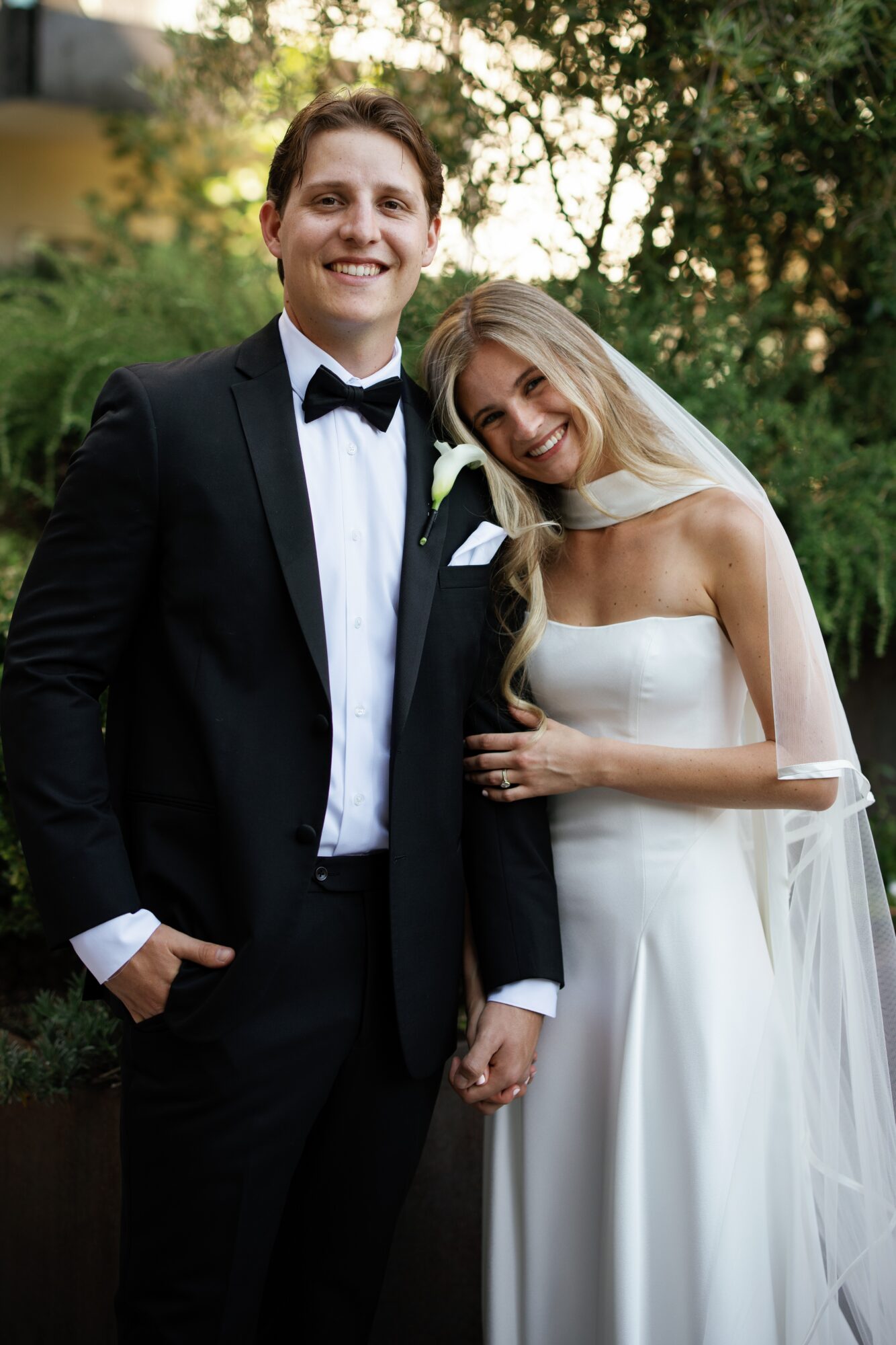 A smiling couple in wedding attire outdoors, holding hands, with trees and sunlight in the background.