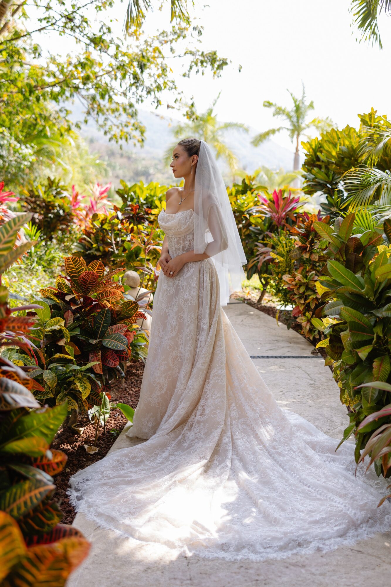 Bride in a white wedding dress and veil standing on a garden path surrounded by colorful plants and trees.