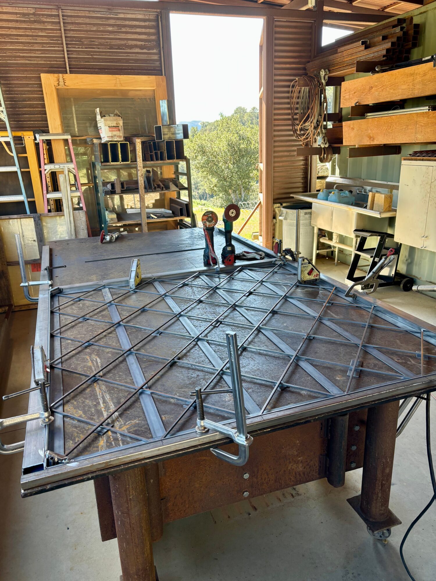 Worktable with metal grid and clamps in a workshop, open door showing trees outside, shelves with tools and materials inside.