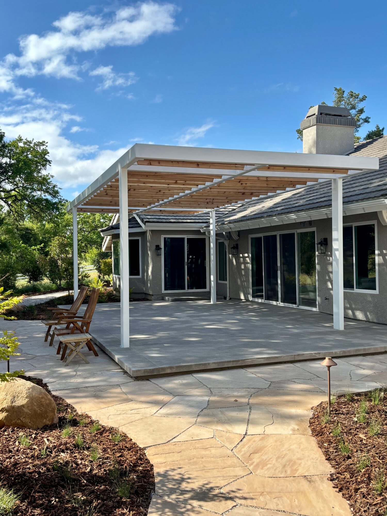 Backyard patio with a white pergola, wooden chairs, and a stone pathway, under a blue sky with clouds.