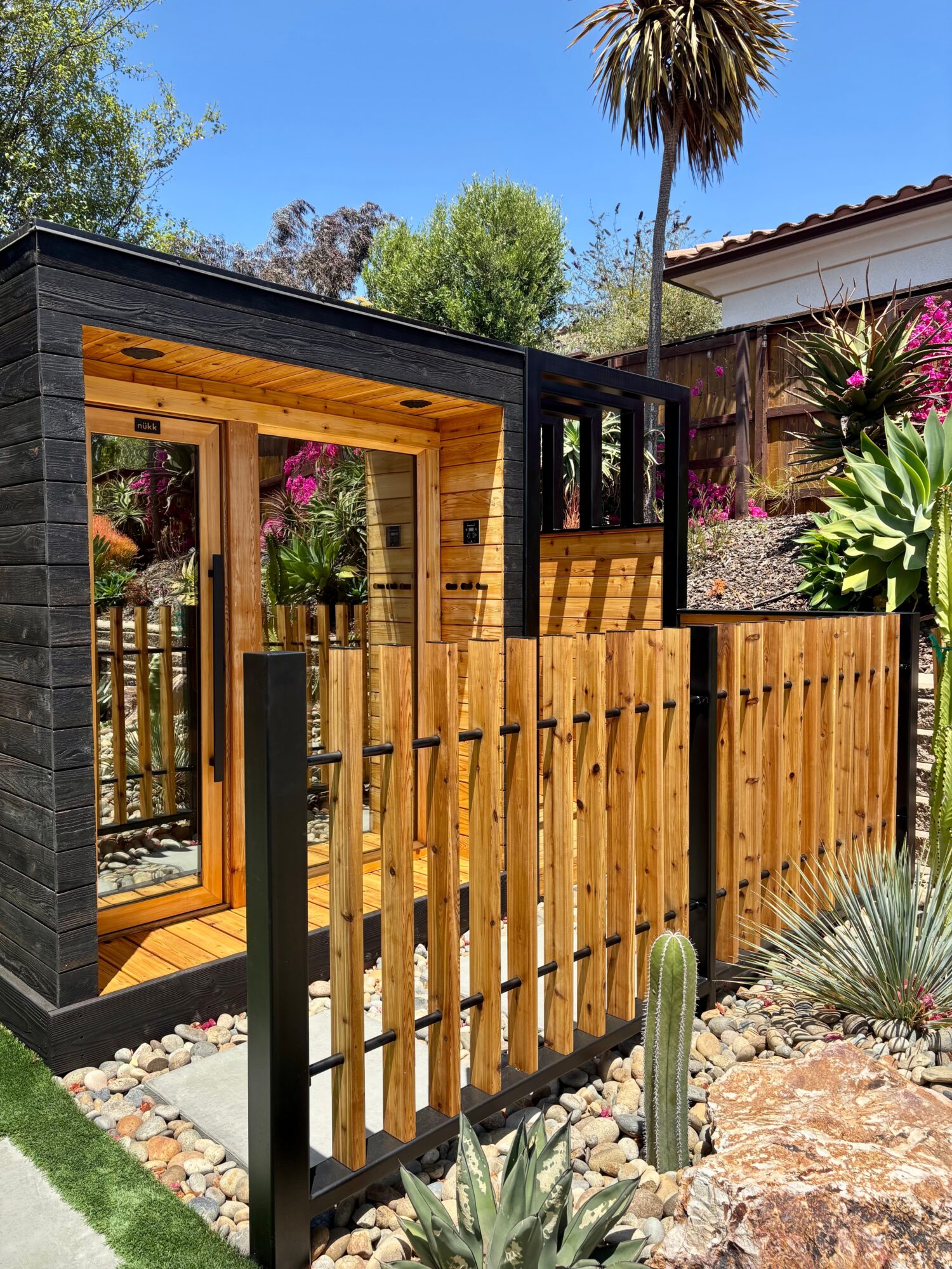 Wooden gate with black frame, garden with plants and trees, blue sky, and a house in the background.