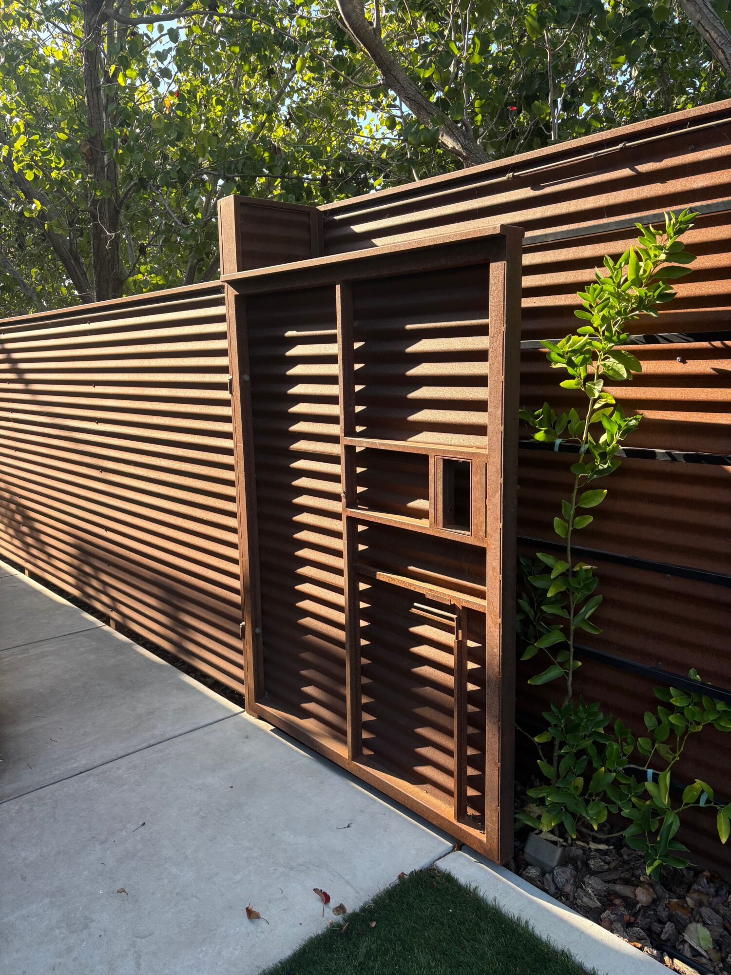 Brown wooden slatted fence with a small door, surrounded by green plants and trees, on a concrete pathway.