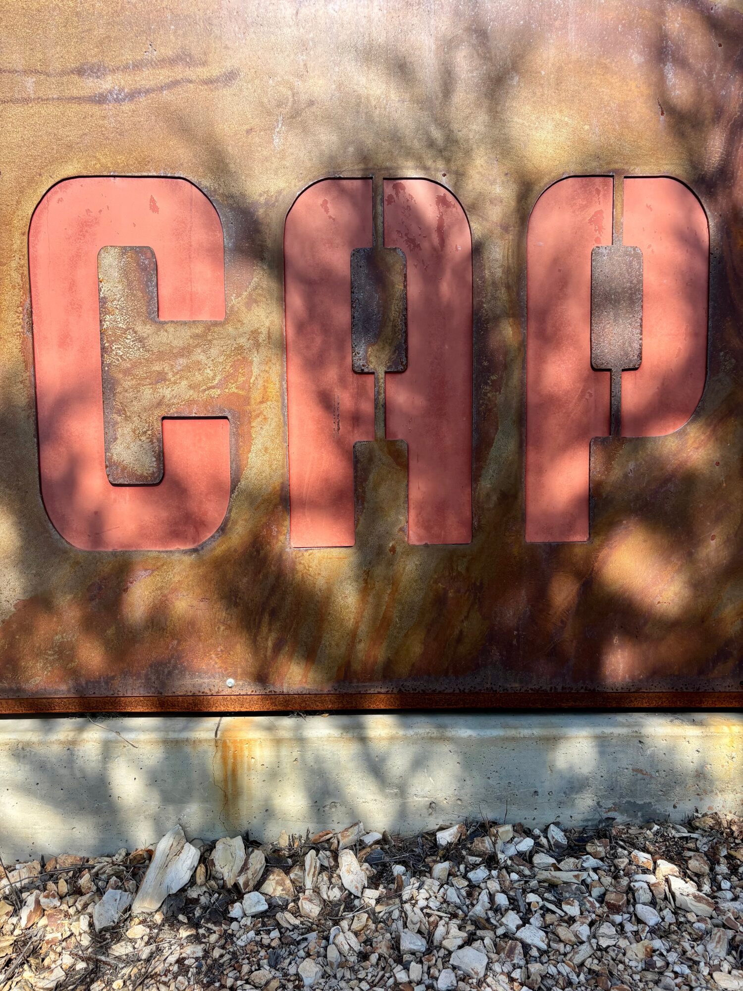 Close-up of a sign with large pink letters spelling 'CAP' on a weathered surface, with shadows and rocks below.