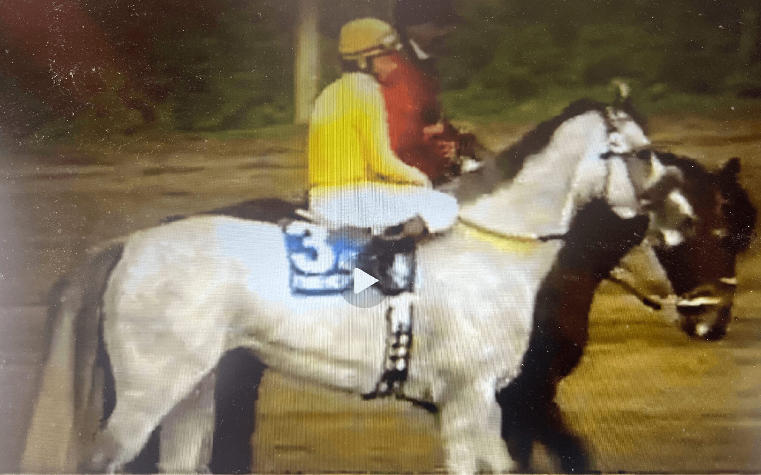 Person riding a white horse with a black mane, wearing a yellow shirt and helmet, on a dirt track.
