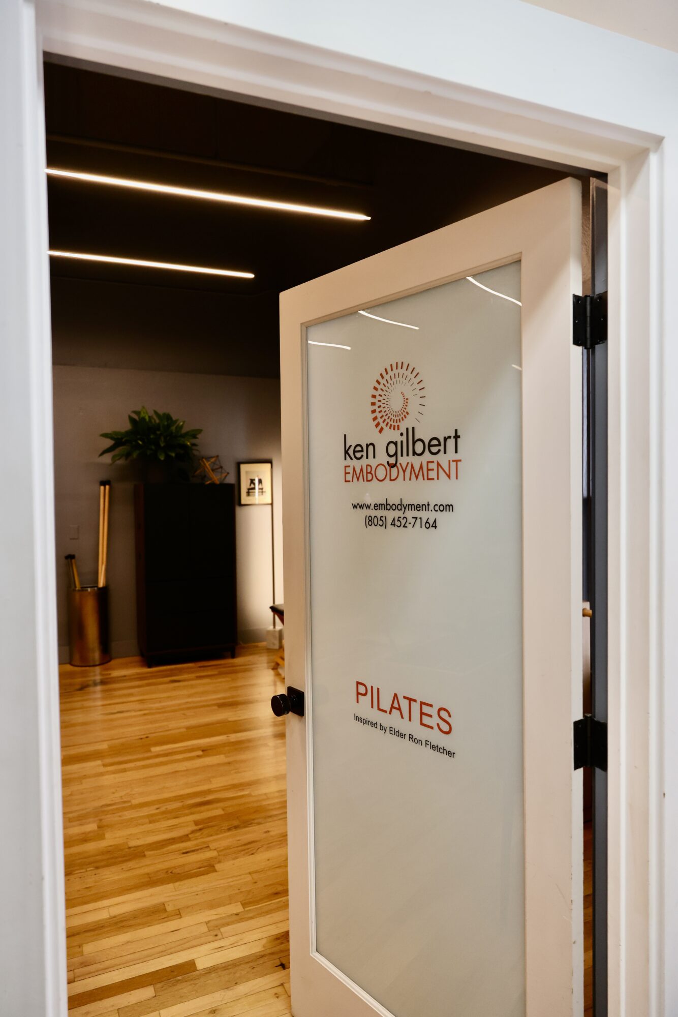 Open door revealing a reception area with wooden flooring, a plant, and ceiling lights, leading to a Pilates studio.