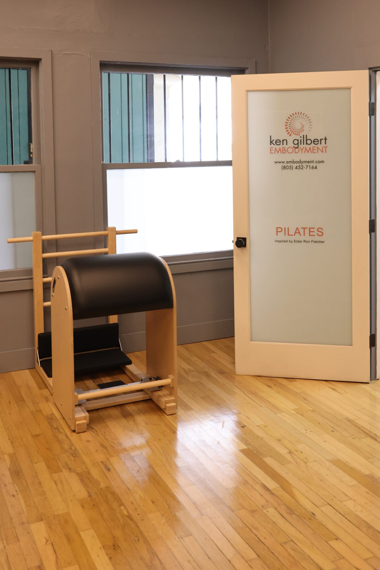 Pilates equipment next to a window with blinds, door with sign, and wooden floor in a studio room.