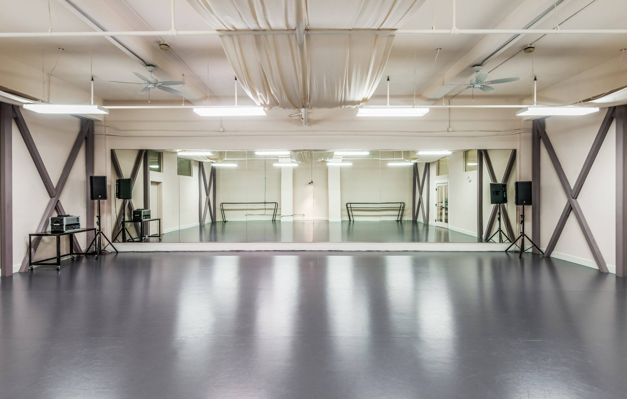 Empty dance studio with a mirrored wall, black floor, and ceiling drapes, with speakers and benches on sides.