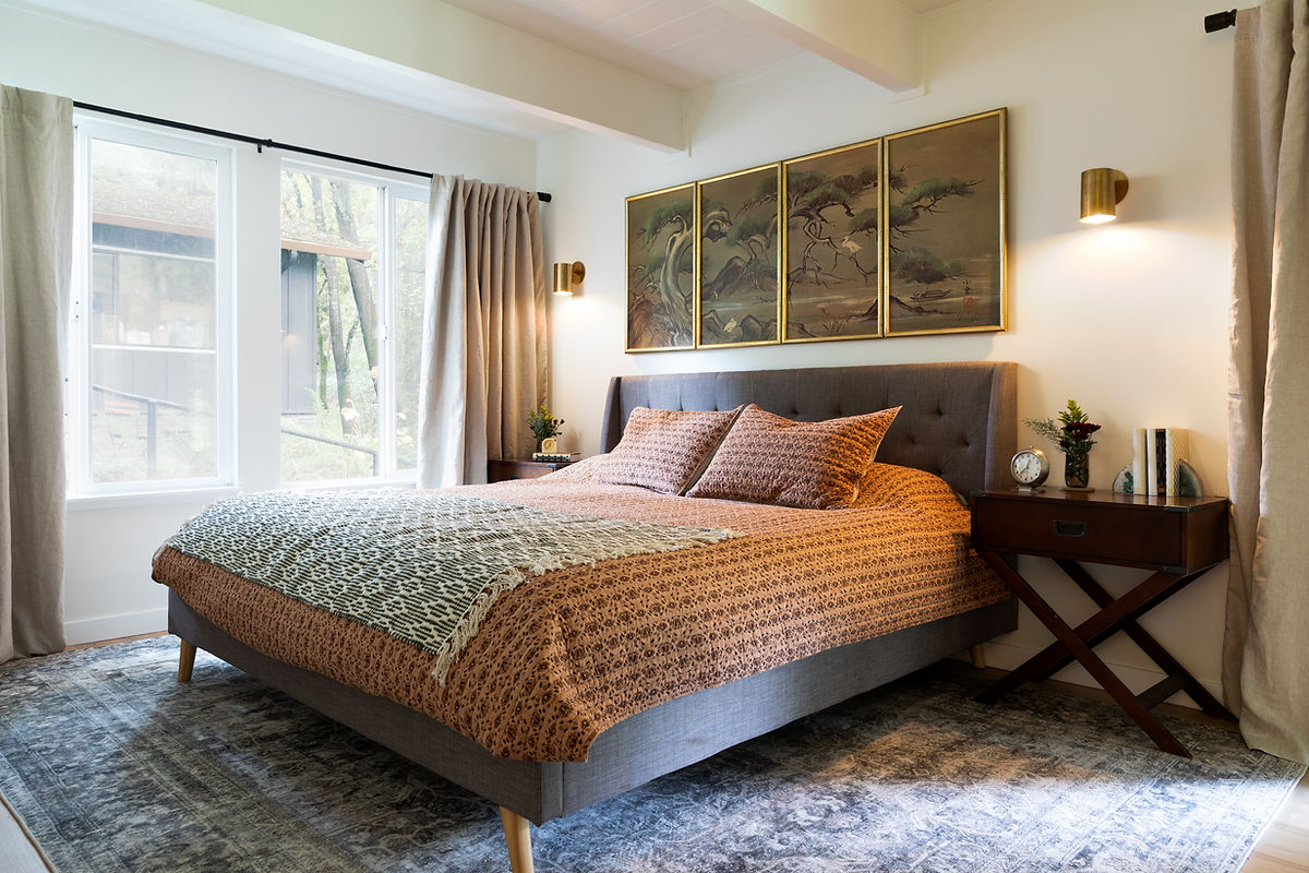 Bedroom with large window, bed with patterned bedding, three-panel landscape artwork above headboard, side tables, and a rug.