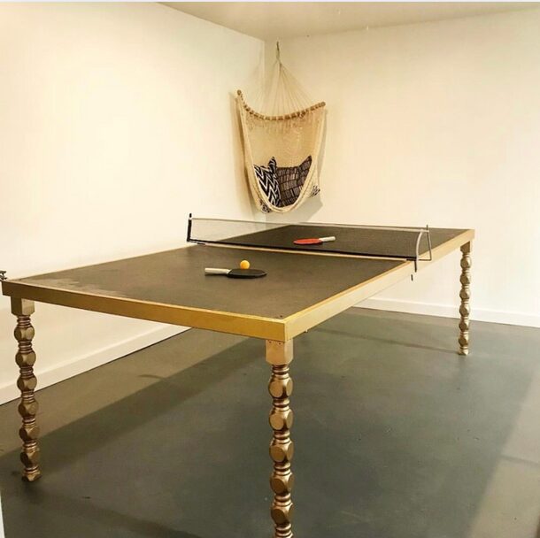 Ping pong table with paddles and balls in a room with a wall hanging and plain walls.
