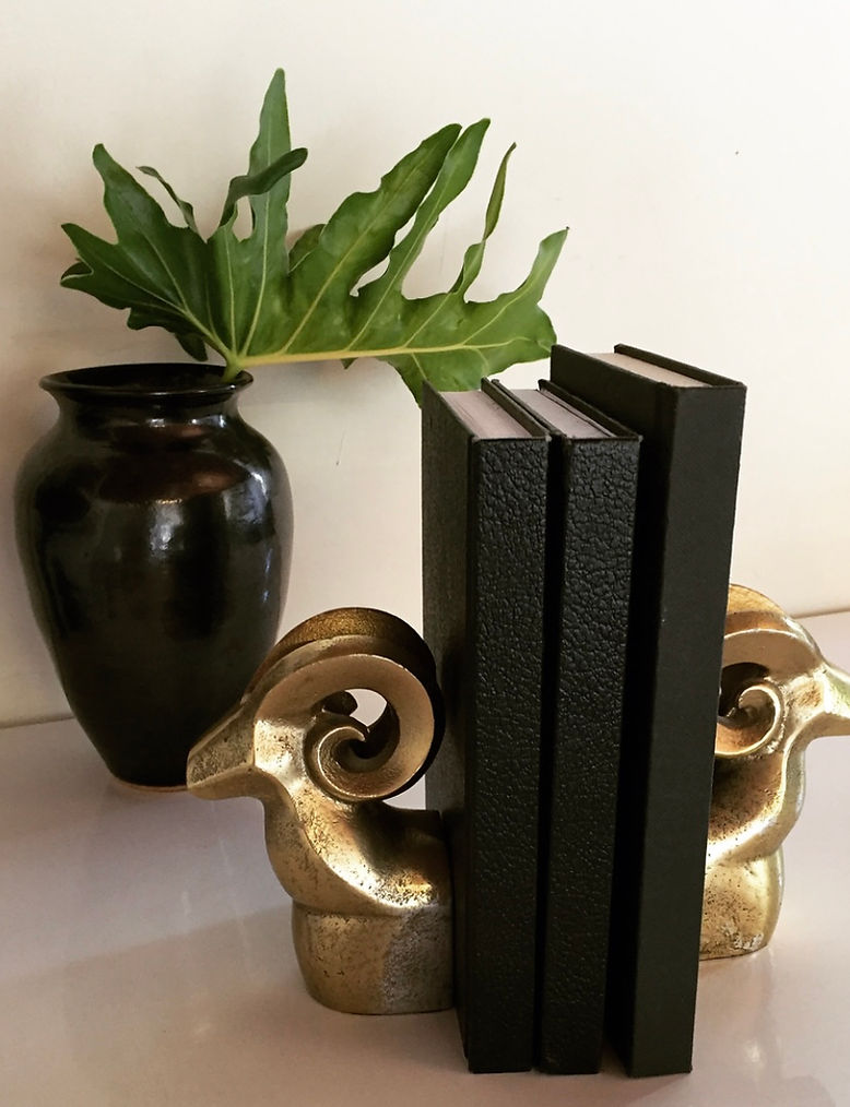 Black vase with green leafy plant, decorative gold and black bookends, and three black books on a white surface.