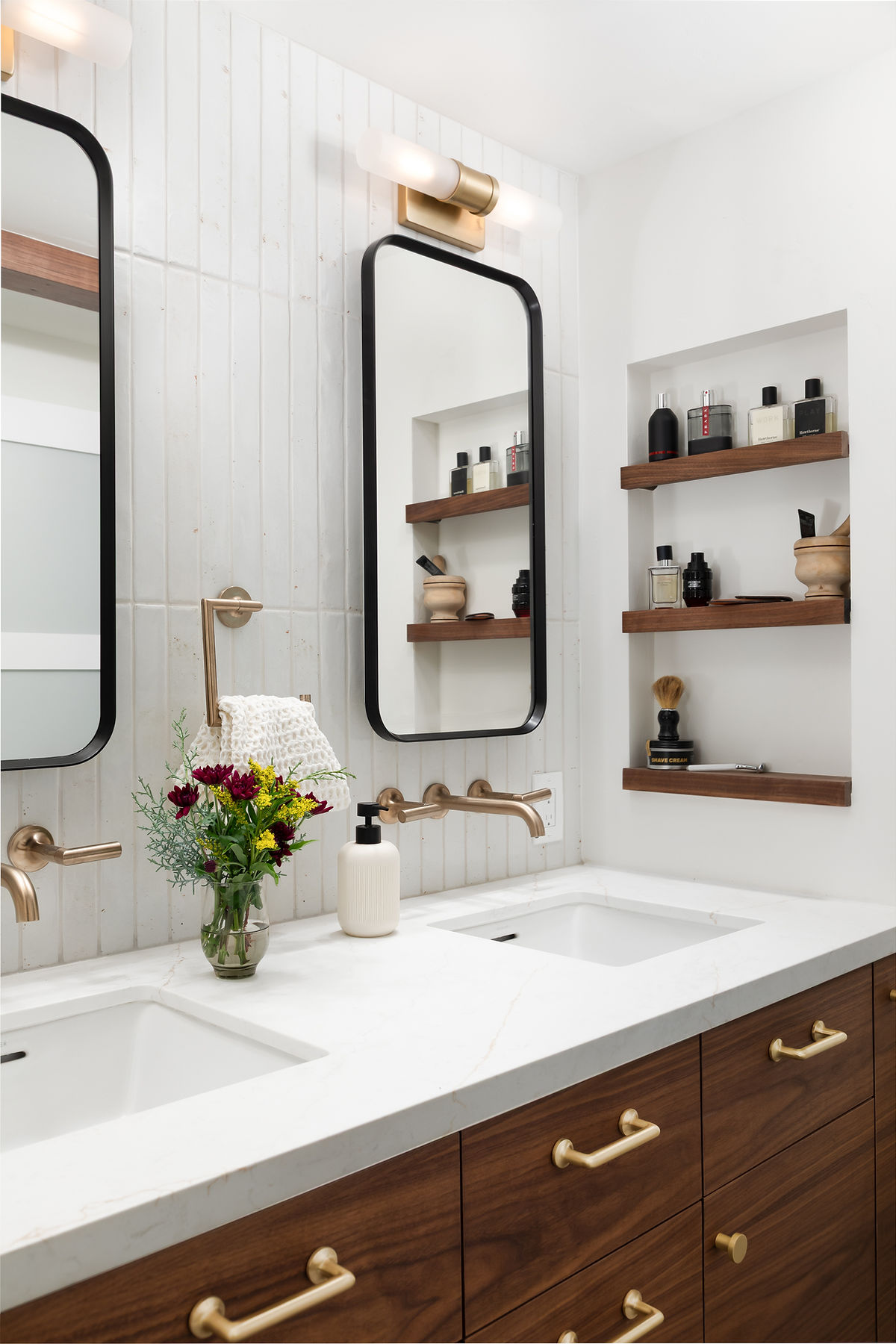 Bathroom vanity with two mirrors, shelves with bottles, a sink, and a vase of flowers, in a bright space.