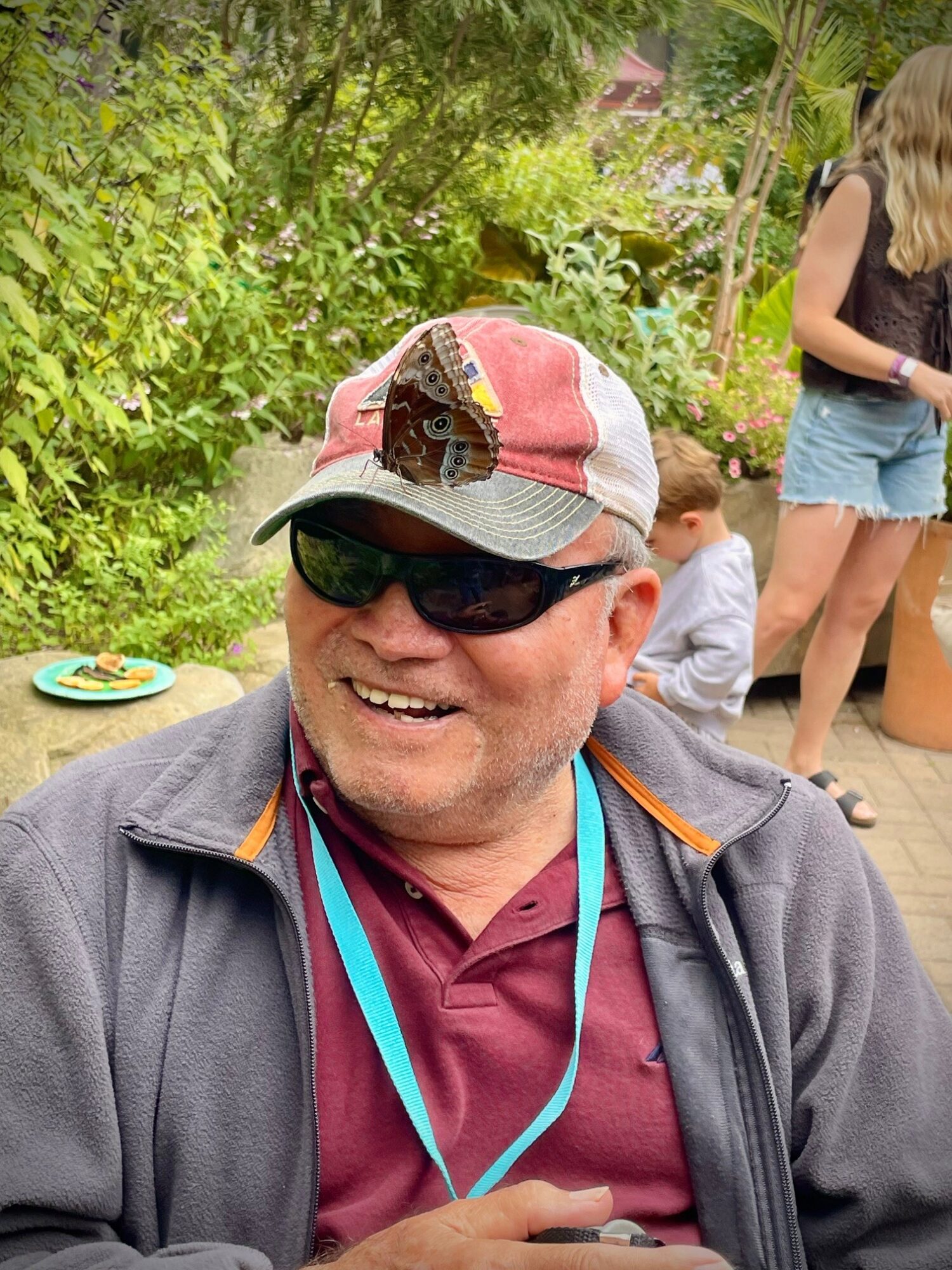 Smiling man wearing sunglasses, a cap, and a jacket outdoors with people and greenery in background.