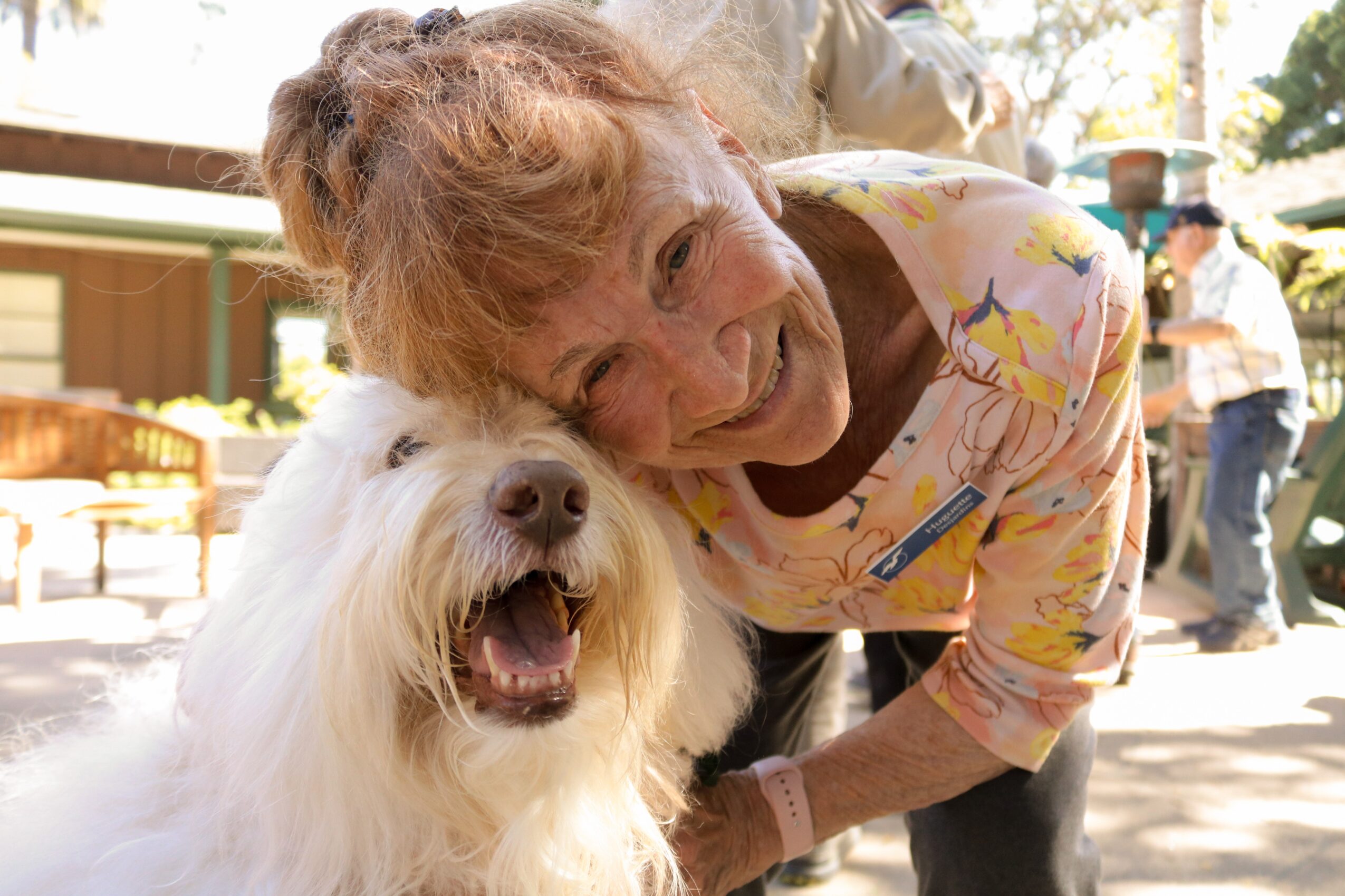 Woman with curly hair smiling and leaning over a fluffy white dog outdoors, sunny day, people in background.
