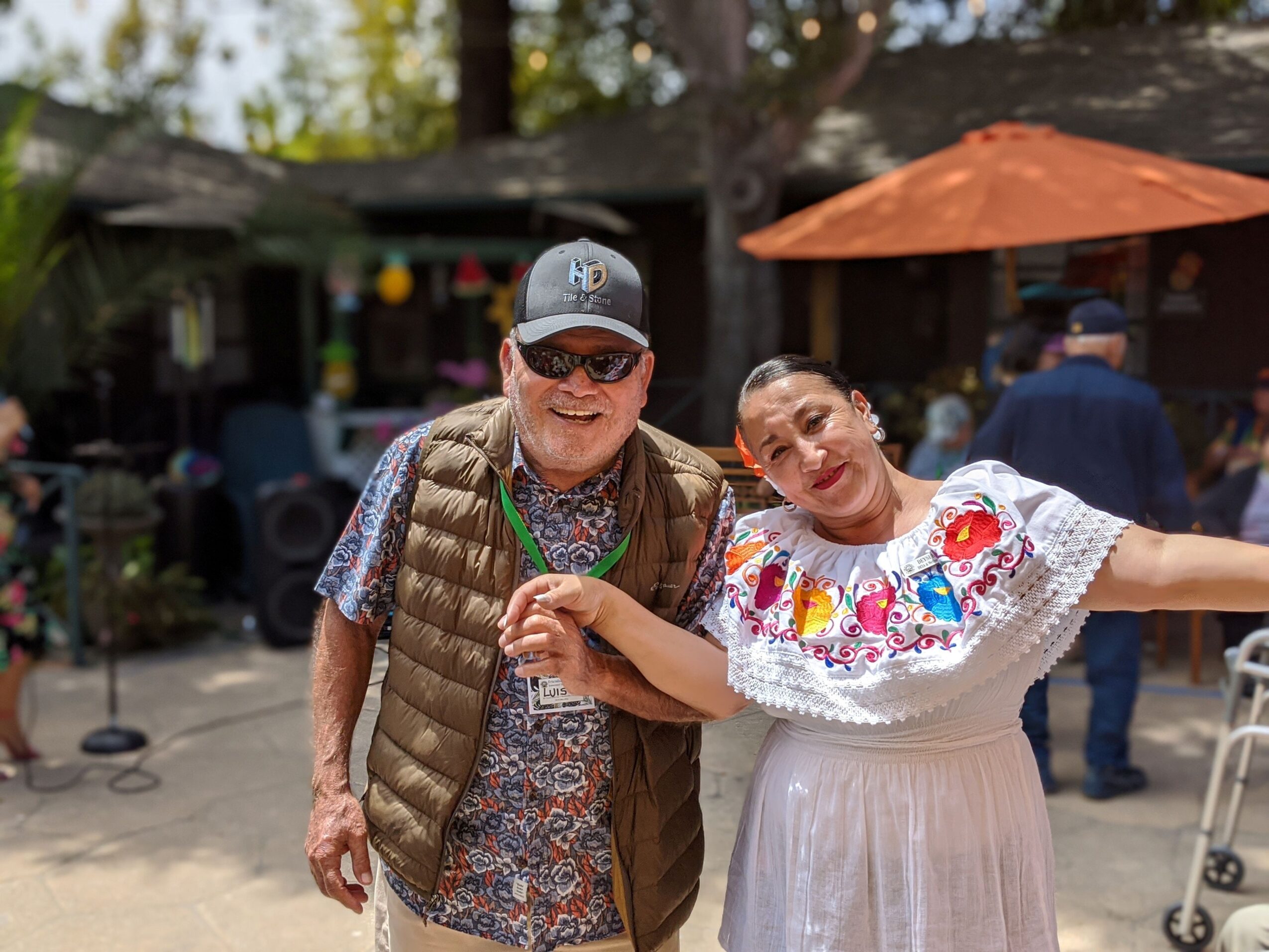 Two people smiling outdoors, one wearing sunglasses and a cap, the other in a white dress with colorful embroidery, holding hands.