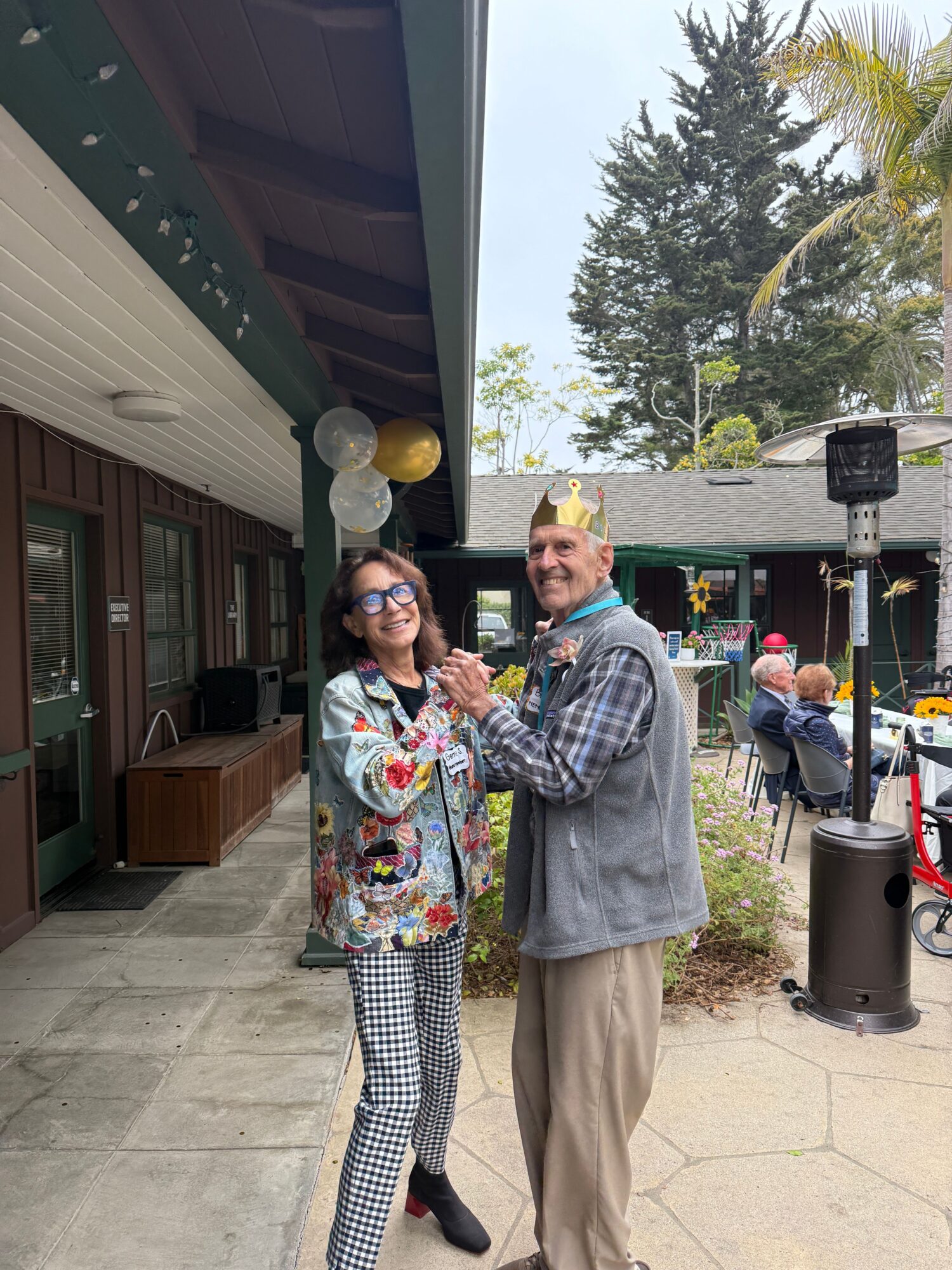 Two people dancing outdoors, one wearing sunglasses and patterned pants, the other wearing a crown and plaid shirt, smiling.