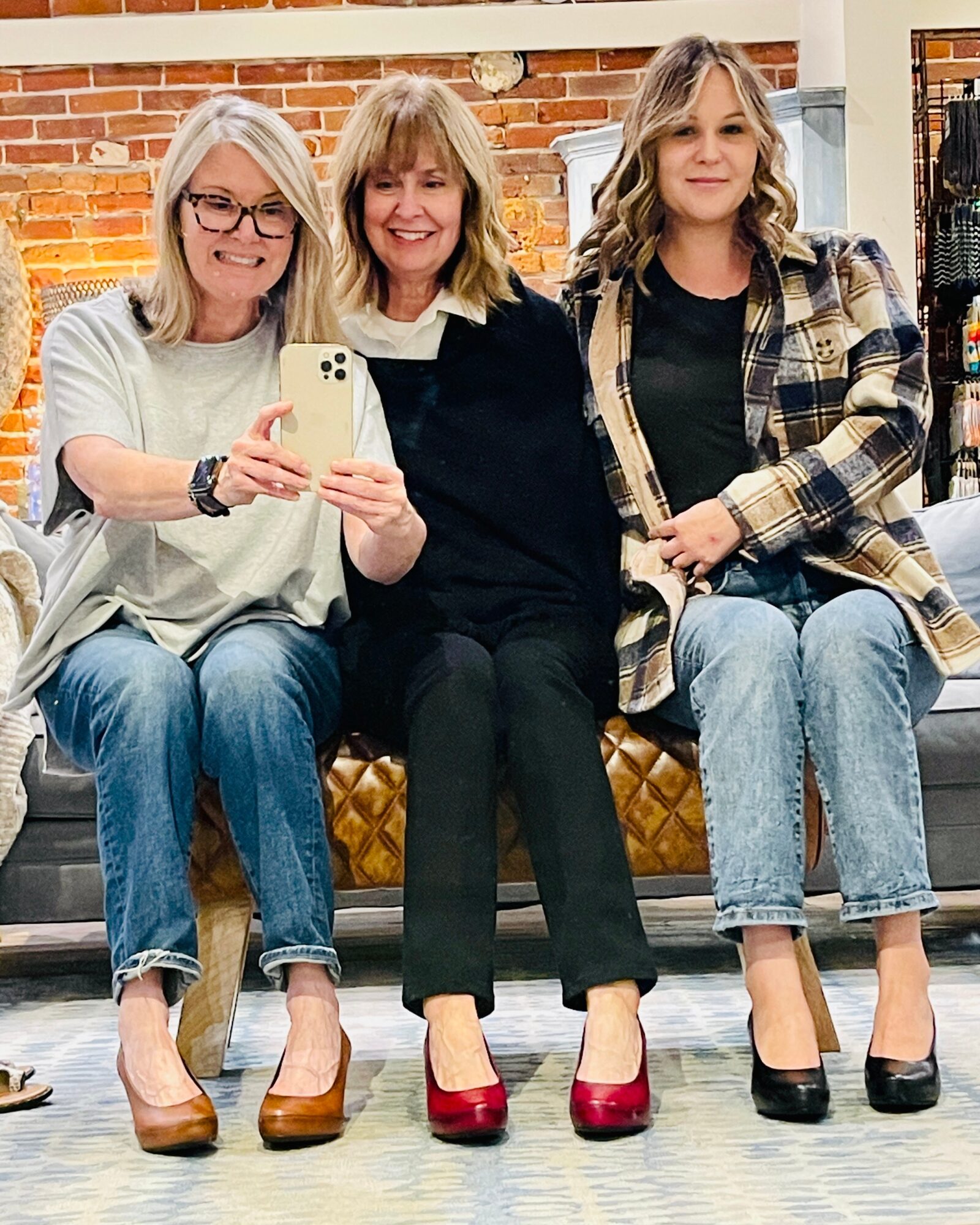 Three women sitting on a bench, smiling, with a brick wall background, taking a selfie. One woman holds a phone, all wear casual clothing.