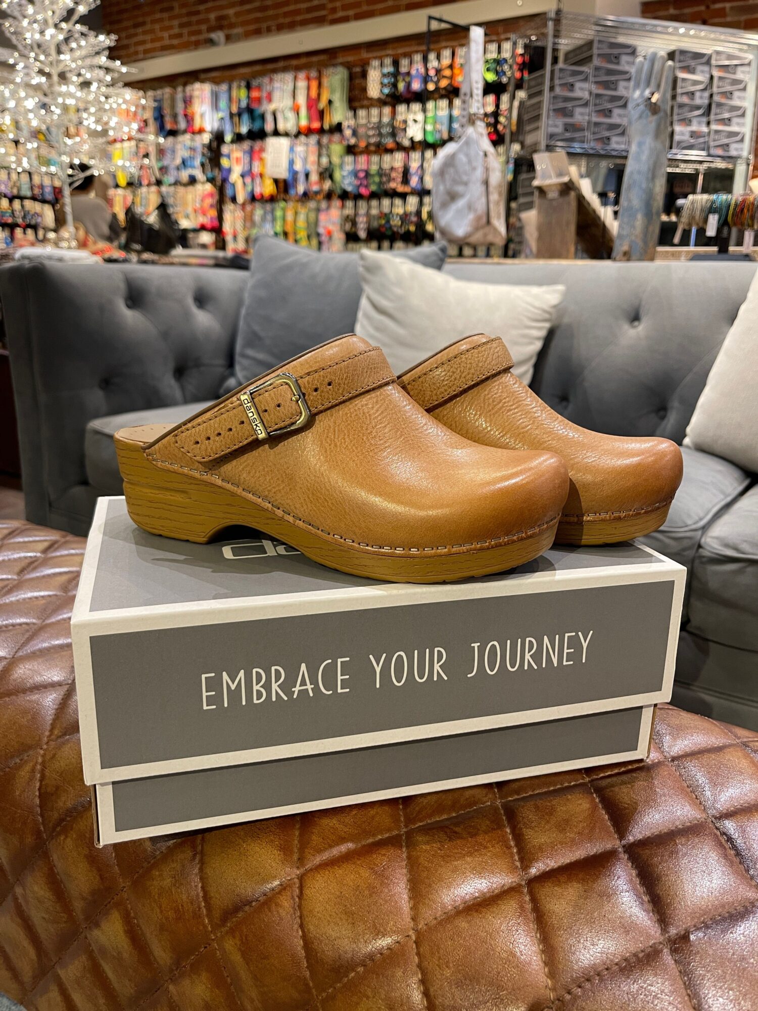 Pair of tan clogs with wooden heels on a display box in a store, with shelves of colorful products in the background.