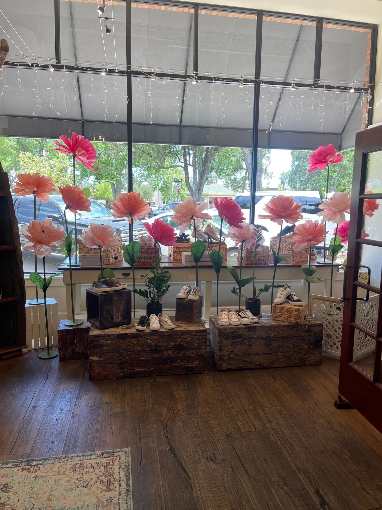 Display of large pink and orange flowers on tall stems in front of a window with trees outside, inside a room.