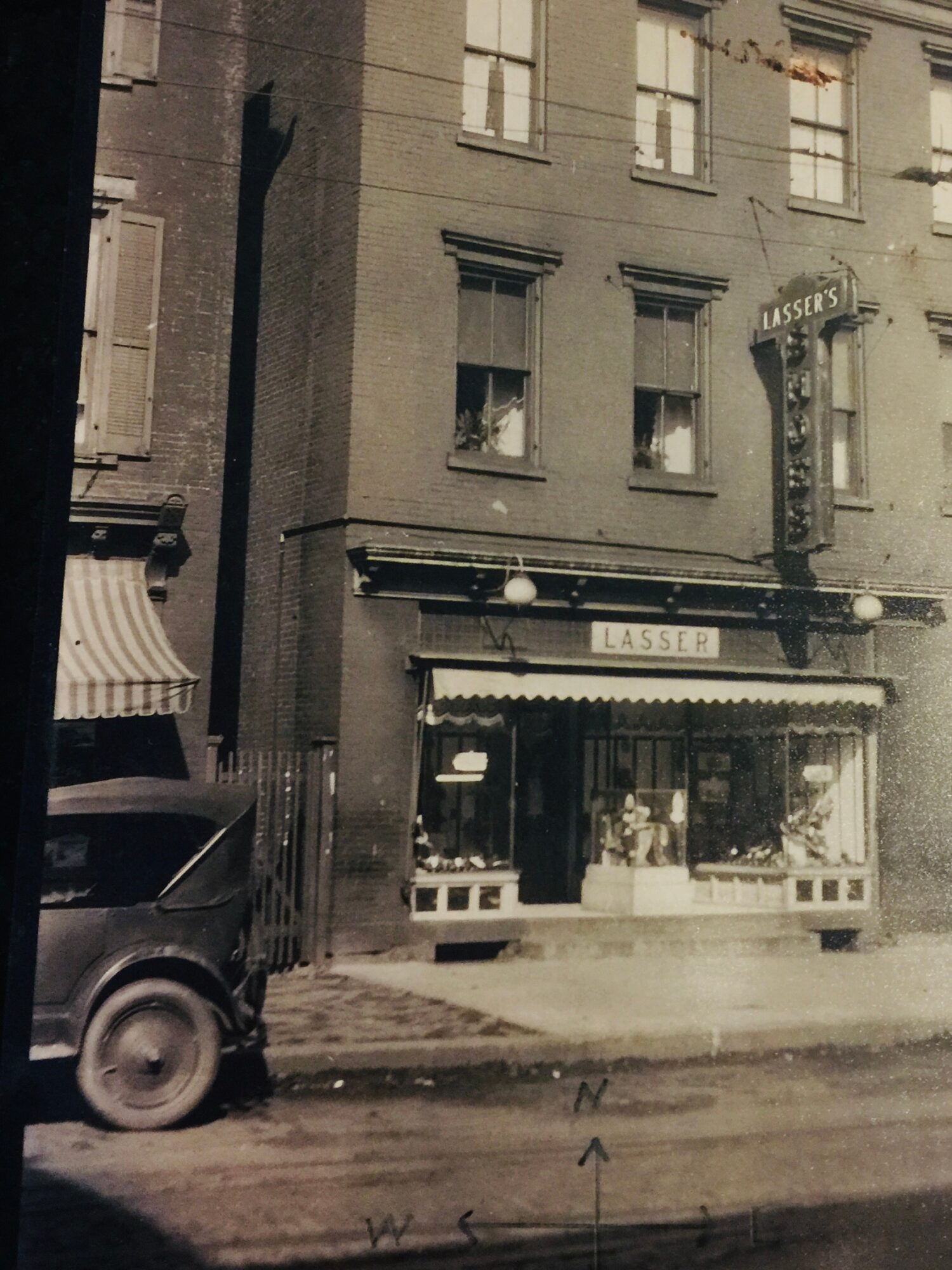 Street view of a three-story building with a shop on the ground floor, awning, and parked vehicle nearby.