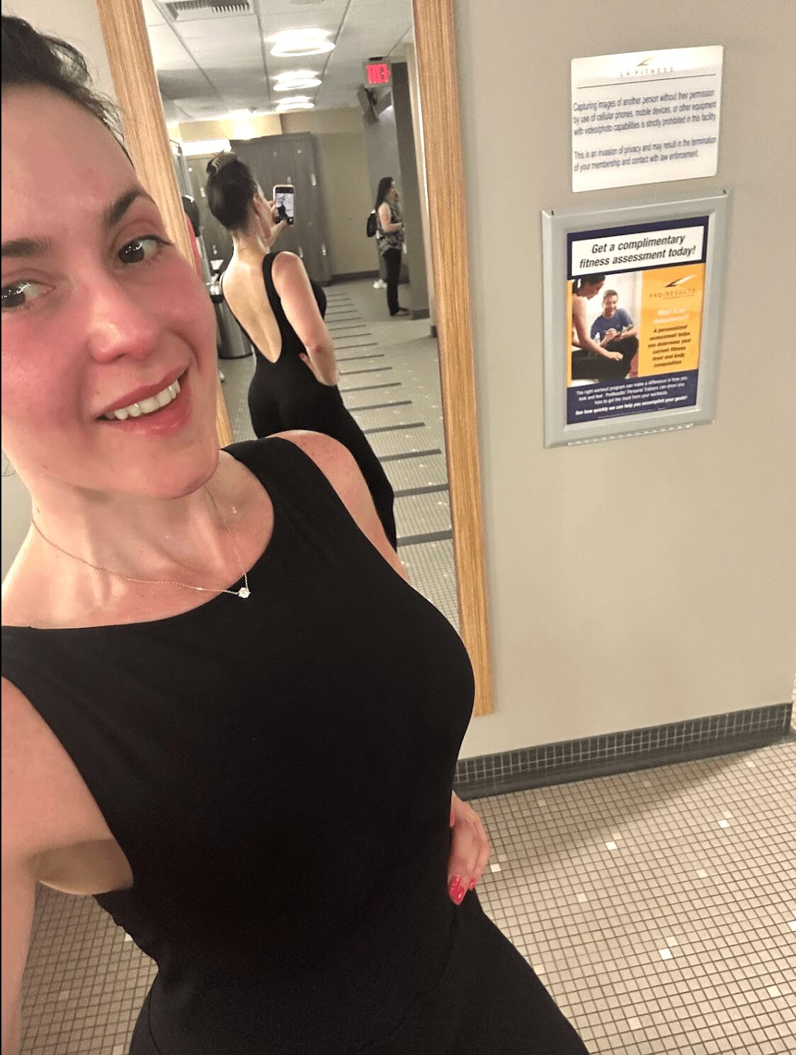 Woman taking a selfie in a mirror, wearing a black sleeveless top, with a bathroom or changing room background.