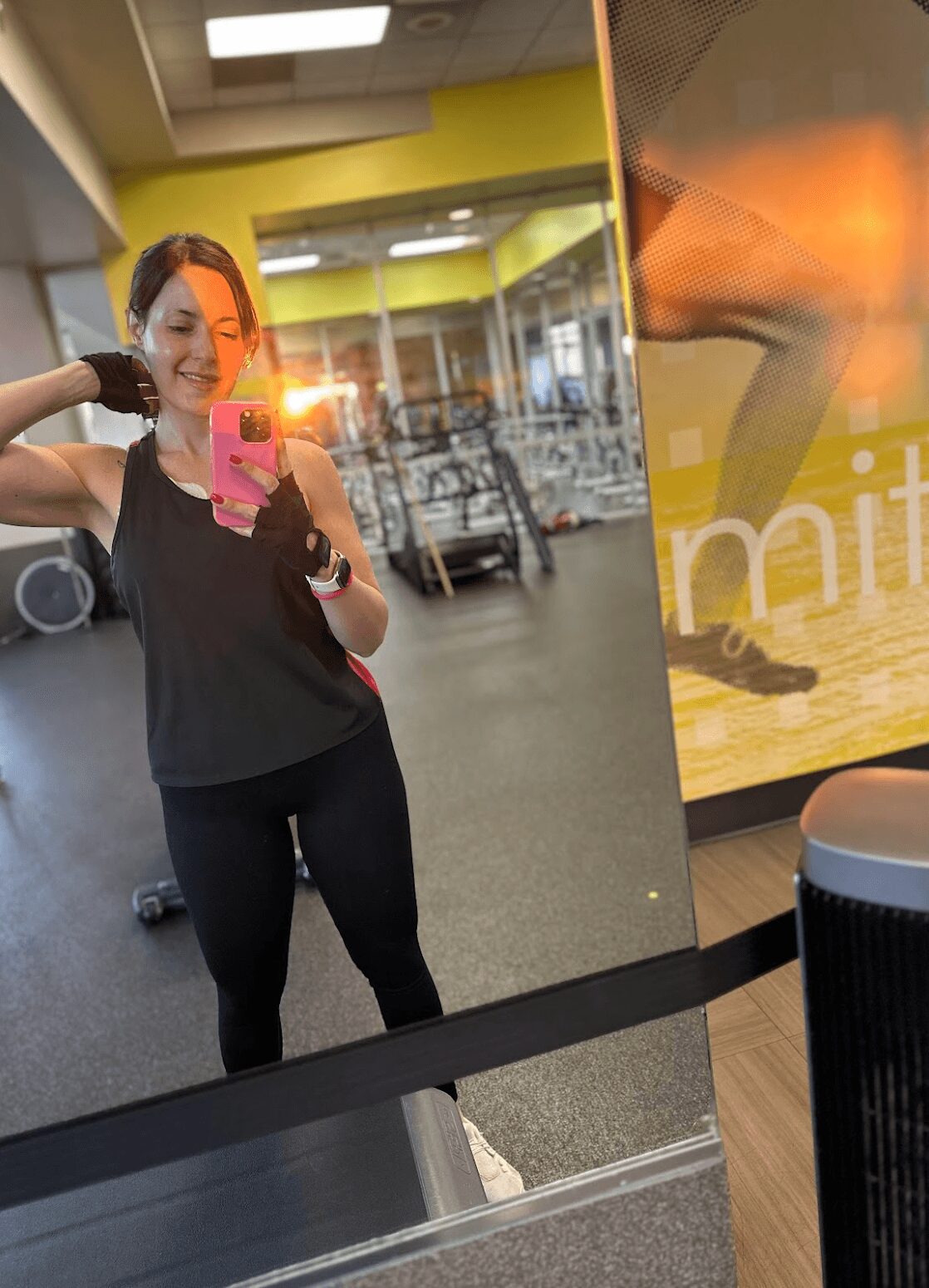 Woman taking a mirror selfie in a gym, wearing black workout clothes, with gym equipment in the background.