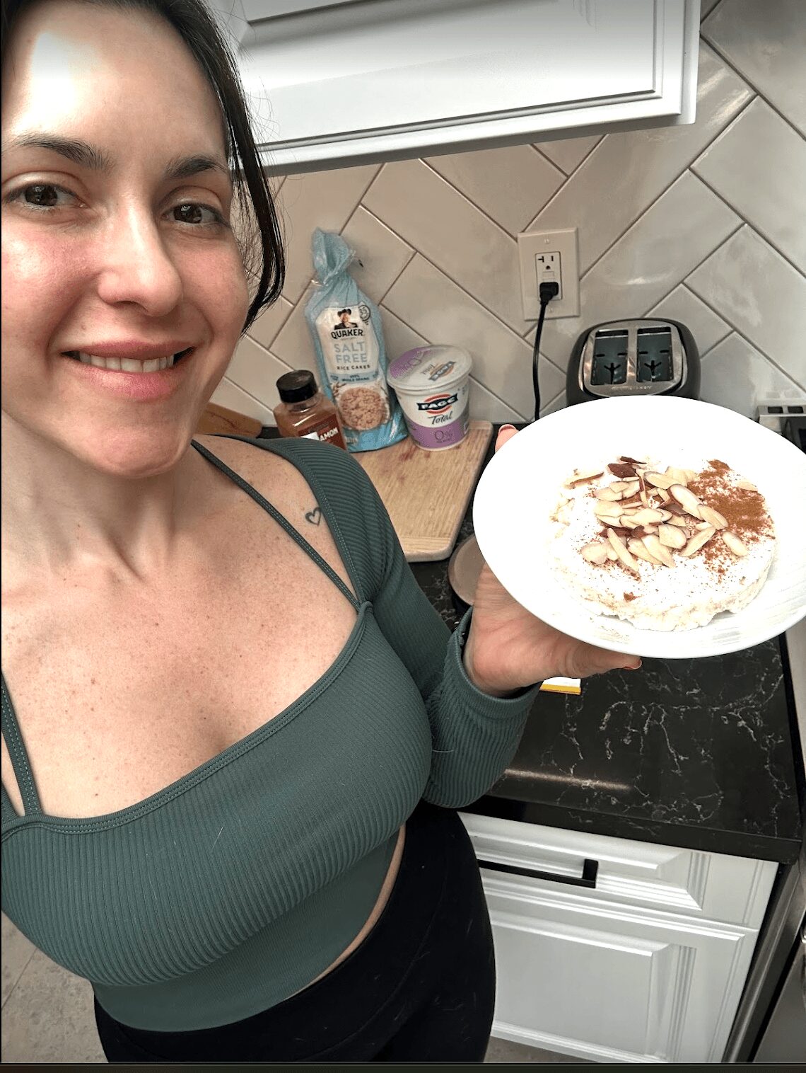 Woman smiling in kitchen holding a plate with sliced almonds and cinnamon, countertop with various items behind her.