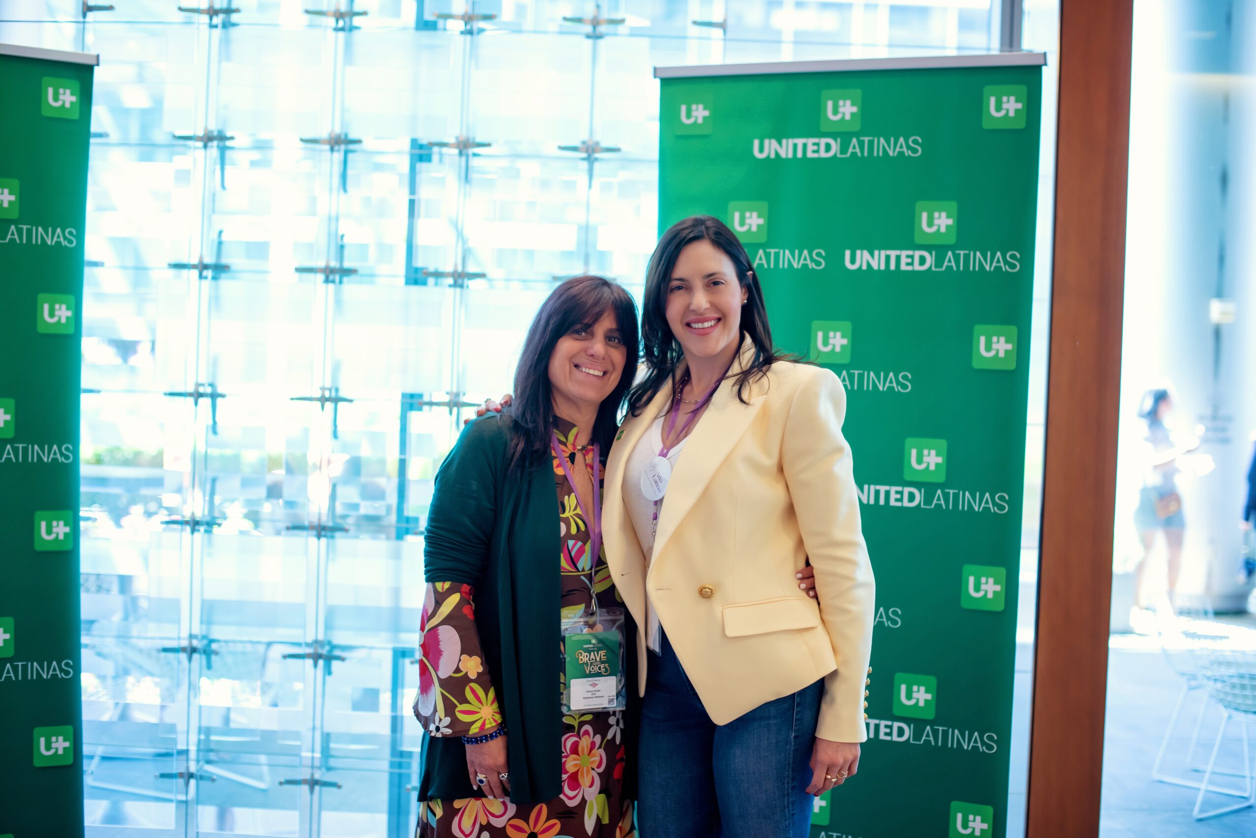 Two women standing together in front of a green backdrop with text, smiling indoors with a glass window behind them.