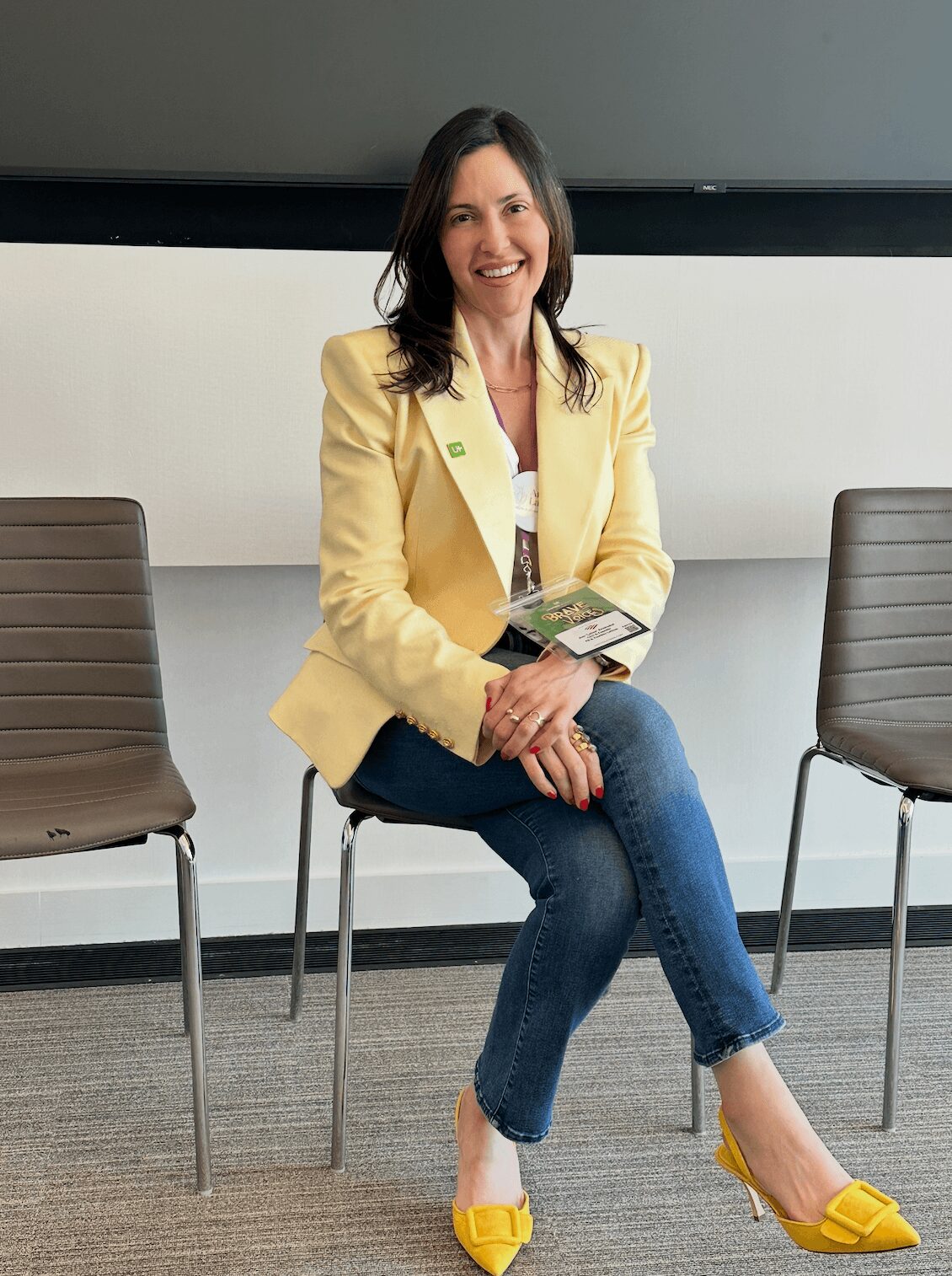 Woman sitting on a chair, smiling, holding a brochure, in a room with two empty chairs, wearing a yellow blazer and jeans.