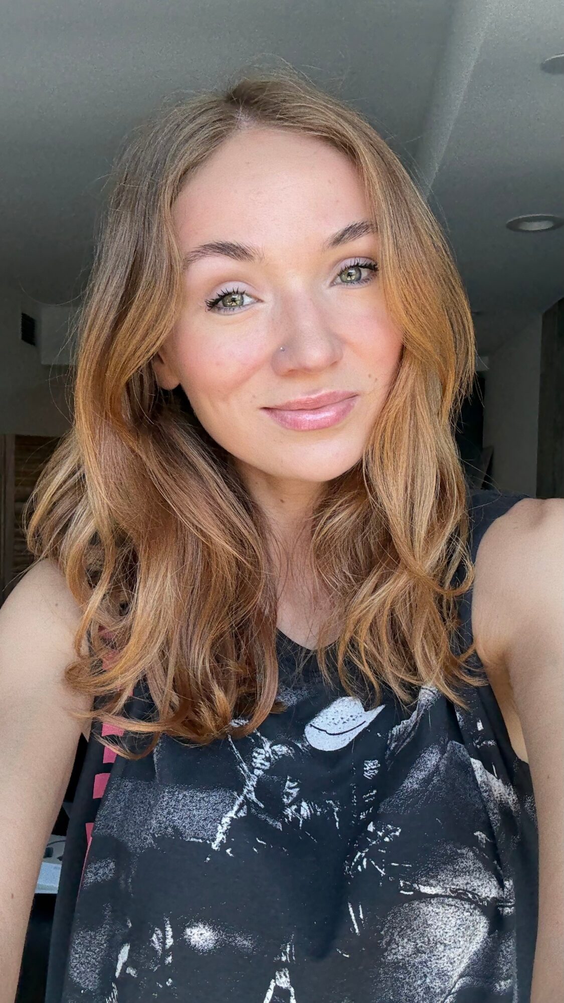 Young woman with wavy, shoulder-length hair smiling, wearing a dark patterned top, indoors with a neutral background.