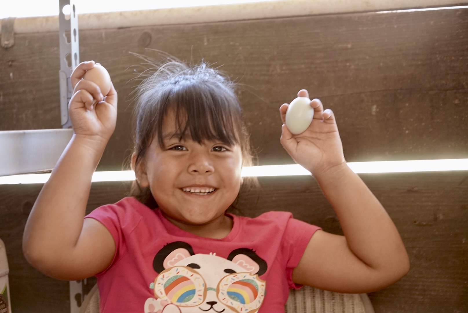 Smiling girl holding an egg in one hand, raising her other arm, wearing a pink shirt with a cartoon character, indoors.