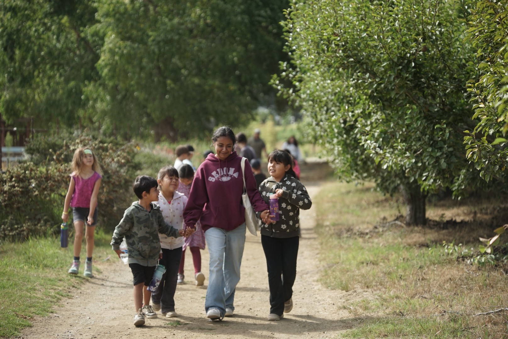 Group of children and an adult walking along a dirt path surrounded by trees on a sunny day.