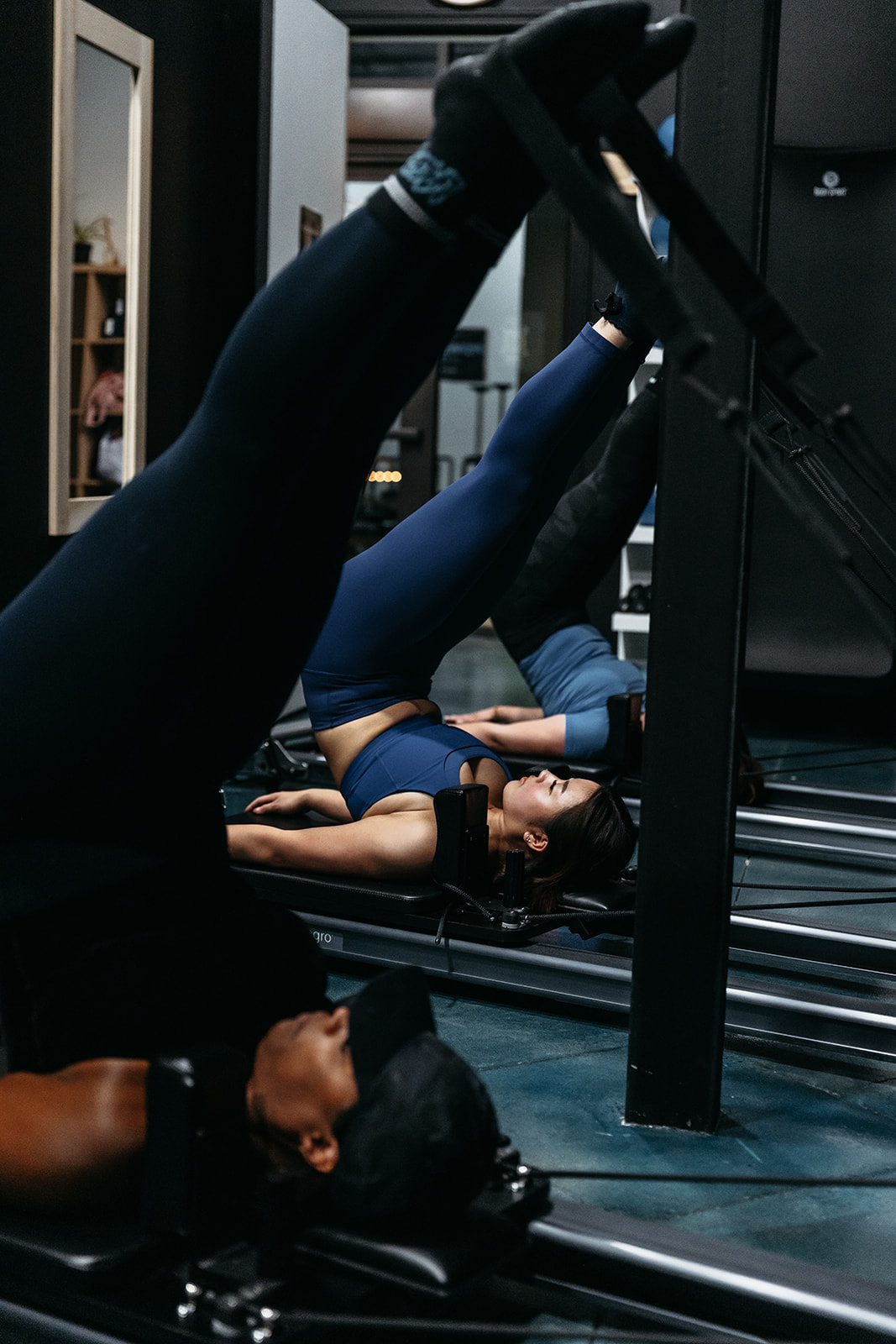 People exercising on gym equipment, lying on their backs with legs raised, in a fitness center.