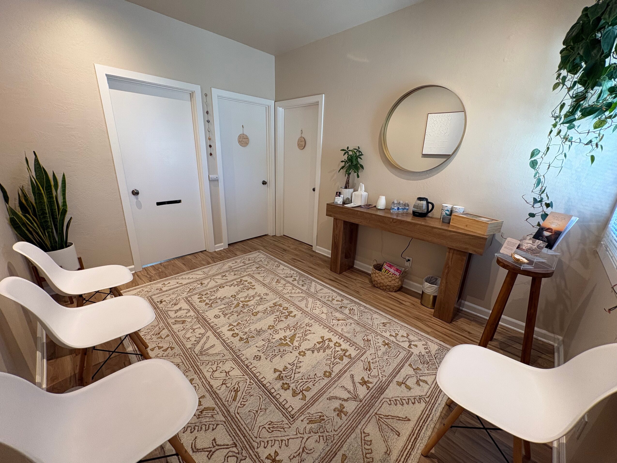 Room with four white chairs, a wooden console table, a round mirror, and a patterned rug on light-colored walls.