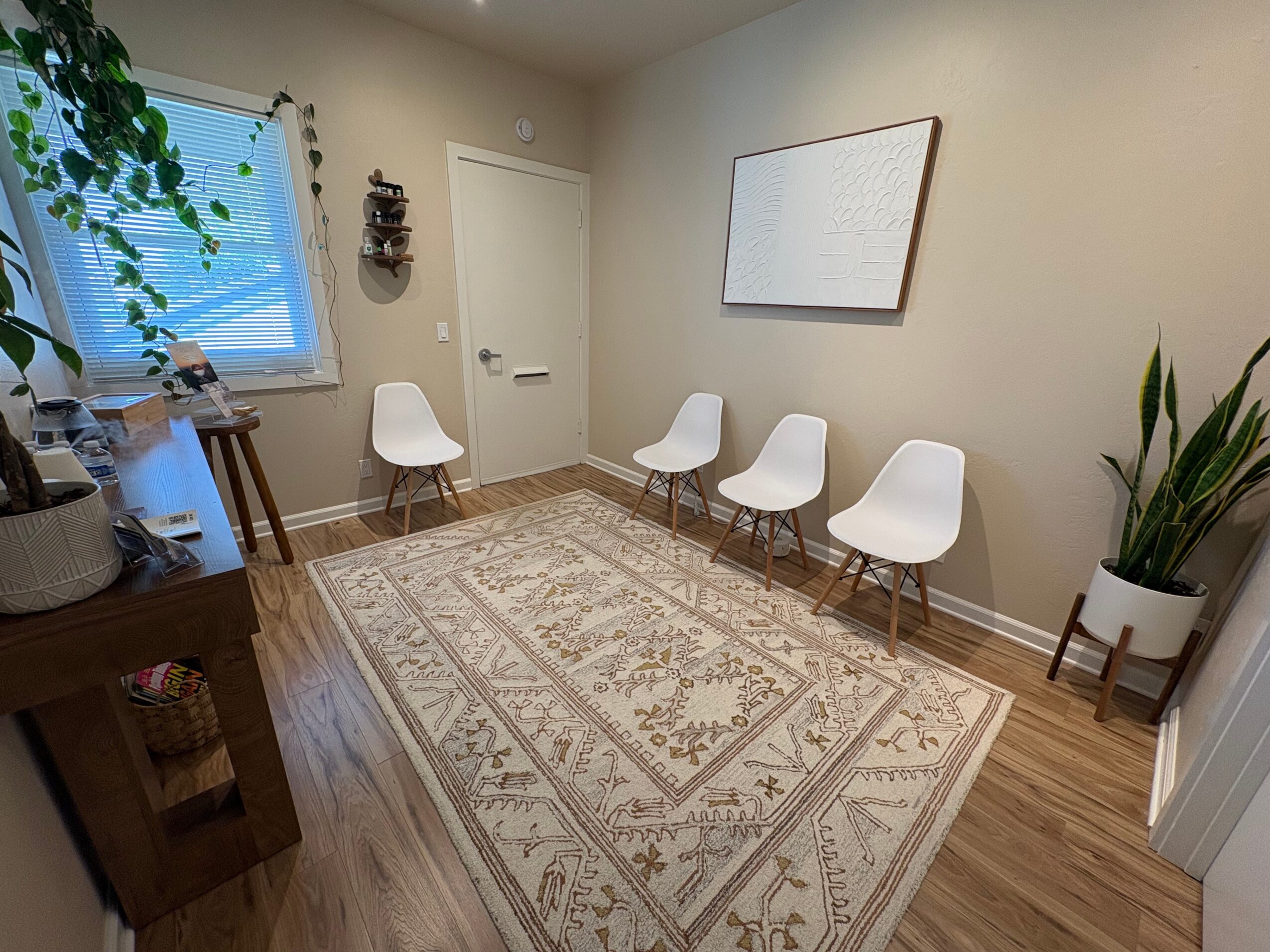 Room with four white chairs, a large patterned rug, a window with blinds, and a potted plant in a white stand.
