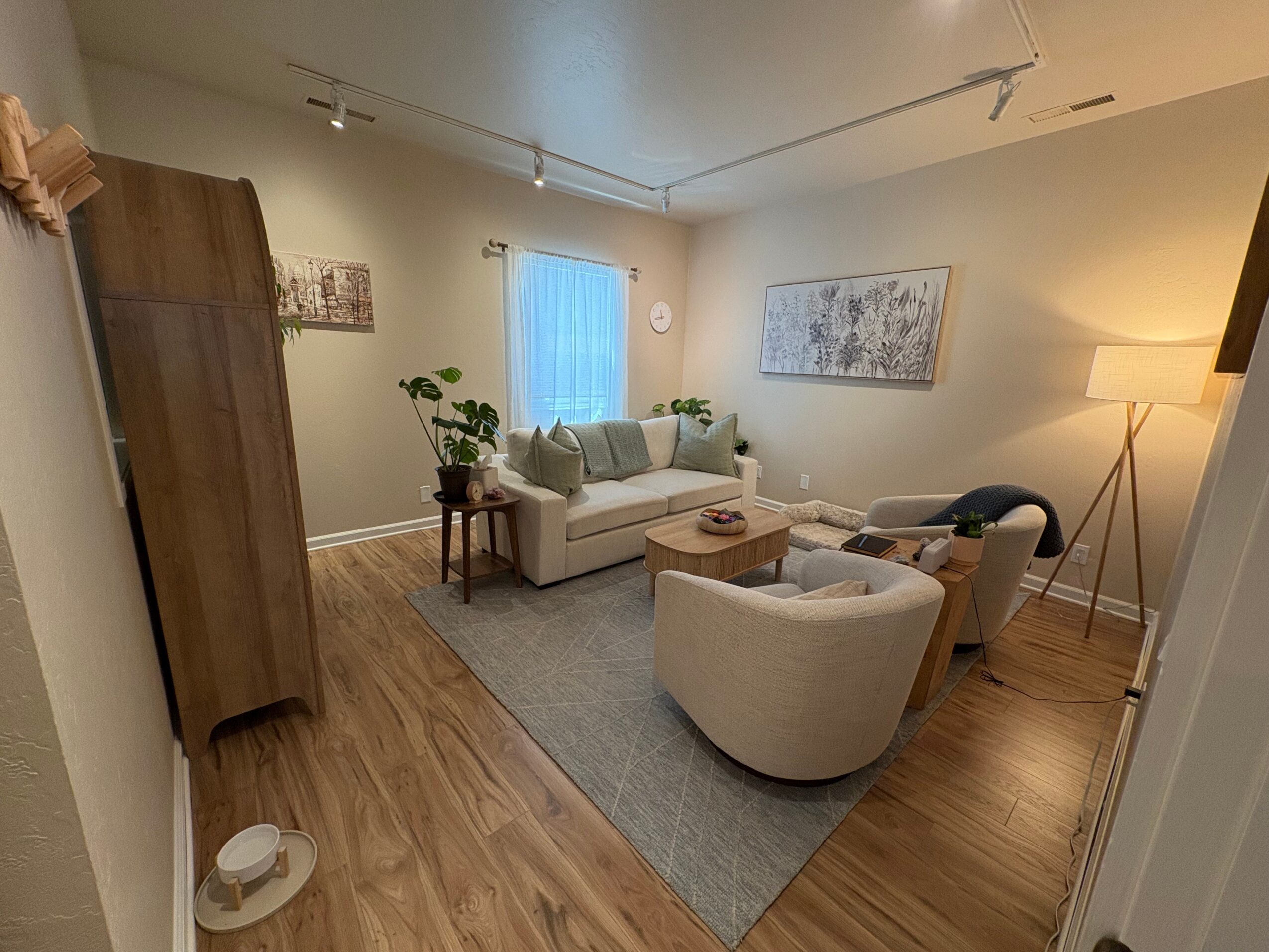 Living room with a white sofa, armchair, wooden coffee table, and a floor lamp, with artwork on the wall and a window with curtains.