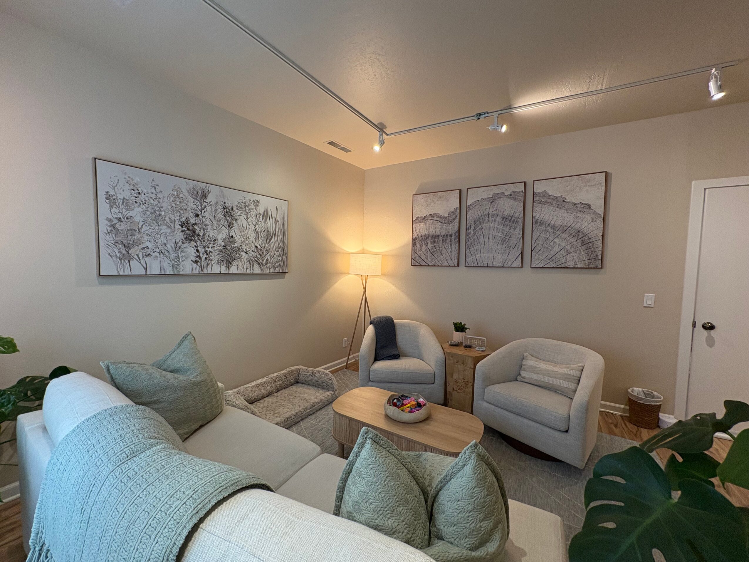 Living room with sofa, armchairs, artwork, and a floor lamp, decorated in neutral tones with plants and a coffee table.
