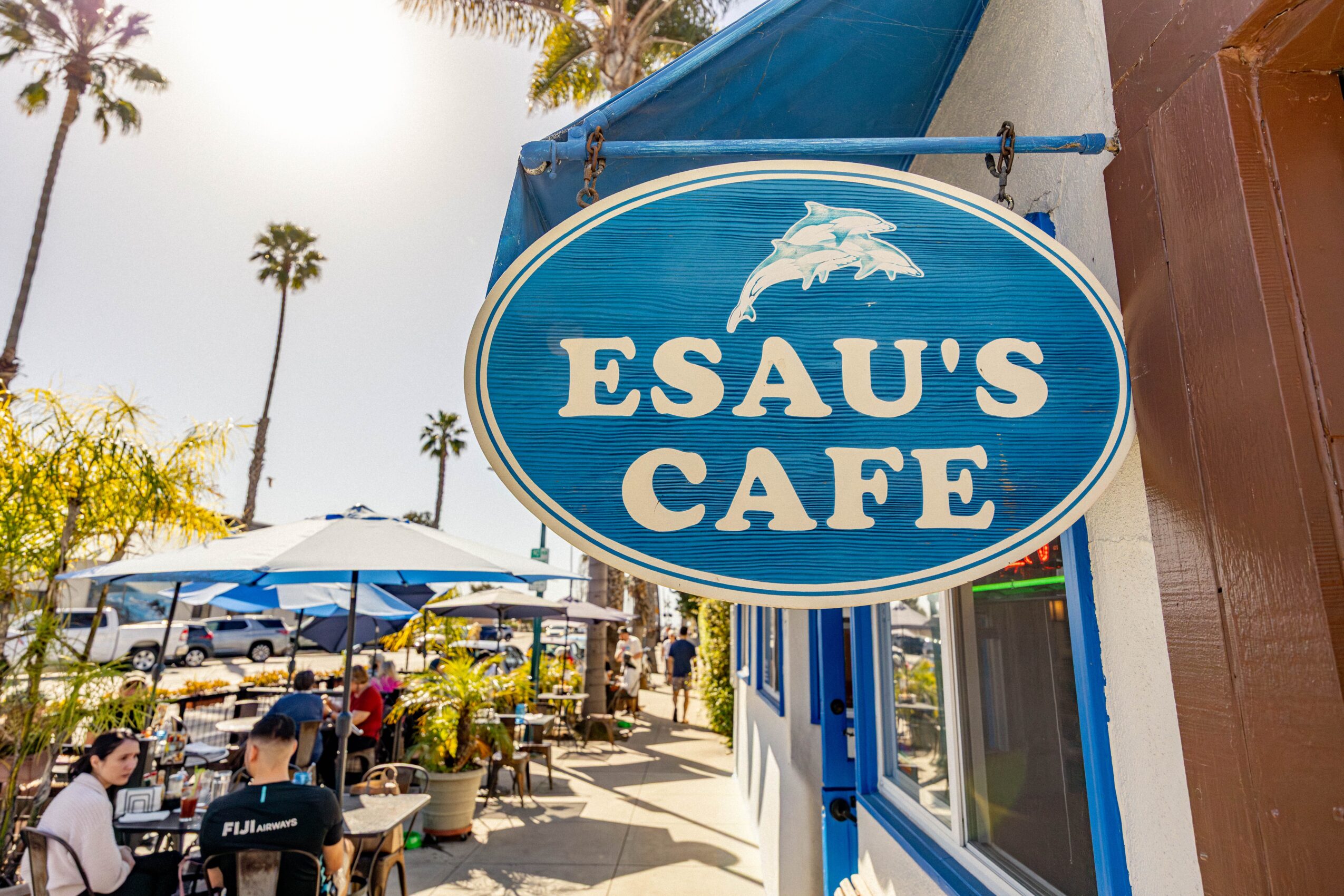 Outdoor cafe with tables, umbrellas, and people, palm trees in background, sunny day, blue sign for Esau's Cafe
