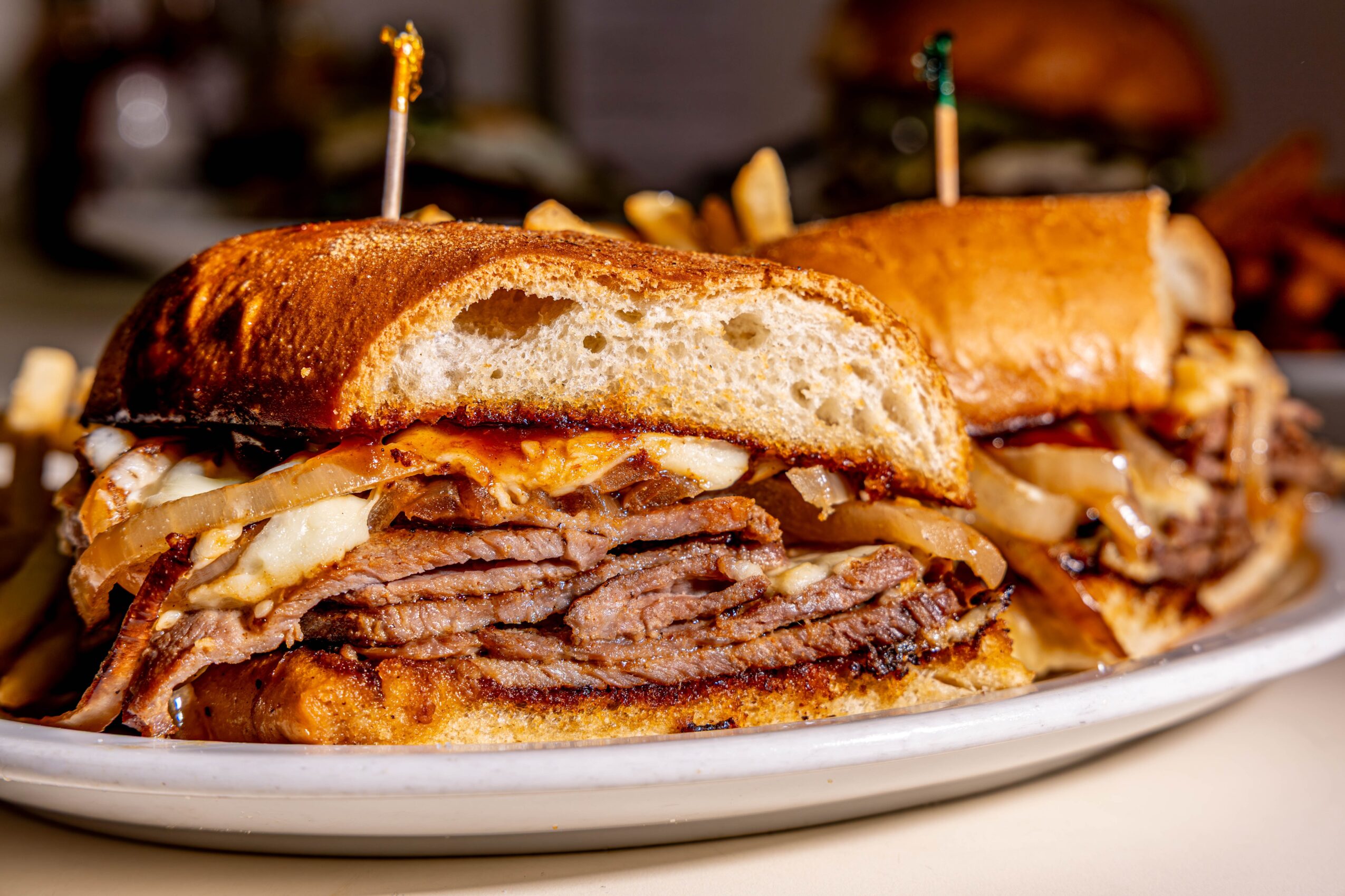 Two large sandwiches with beef, onions, and bread on a white plate, with toothpicks holding them together.