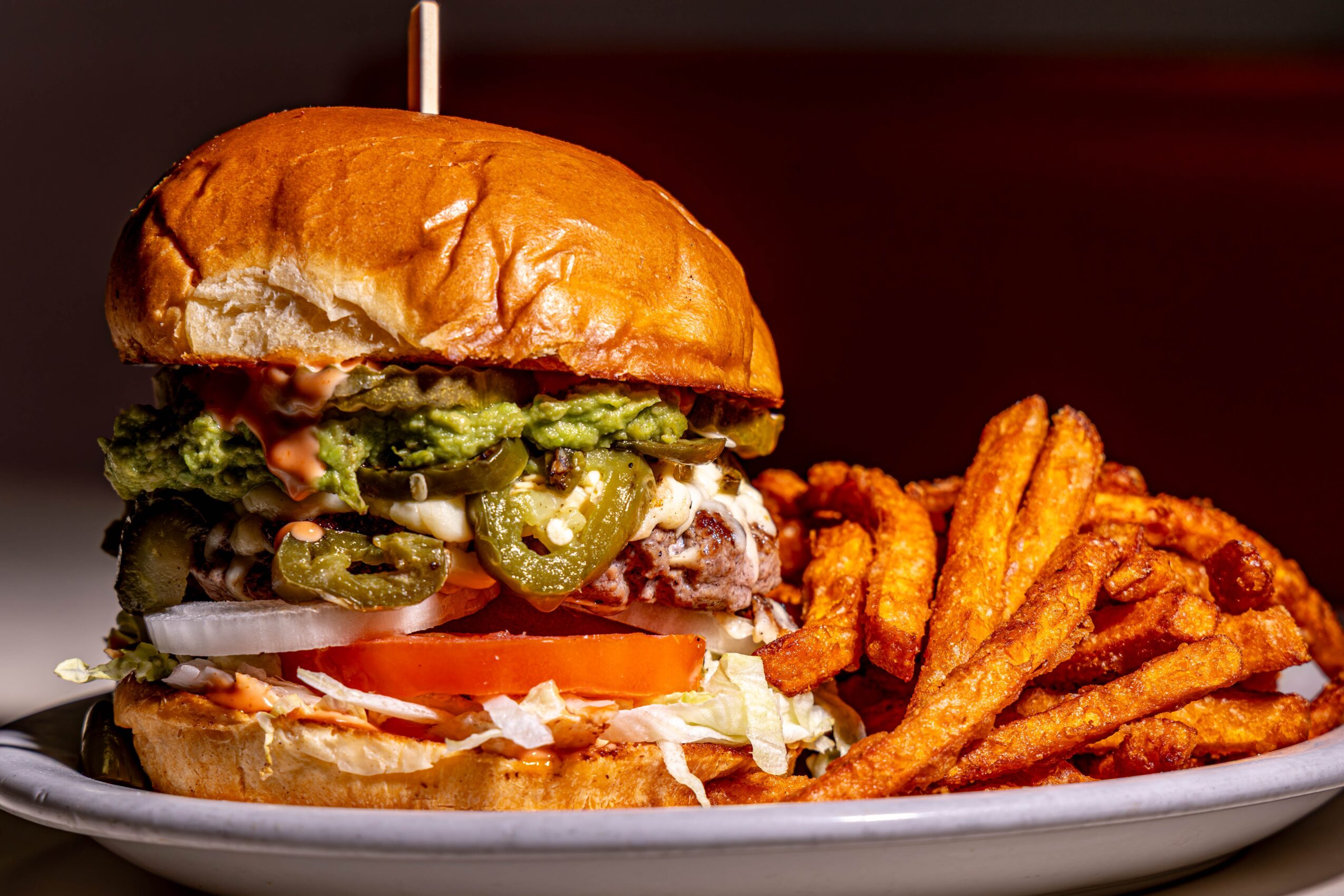 A burger with lettuce, tomato, pickles, onions, and a bun, served with curly fries on a white plate.