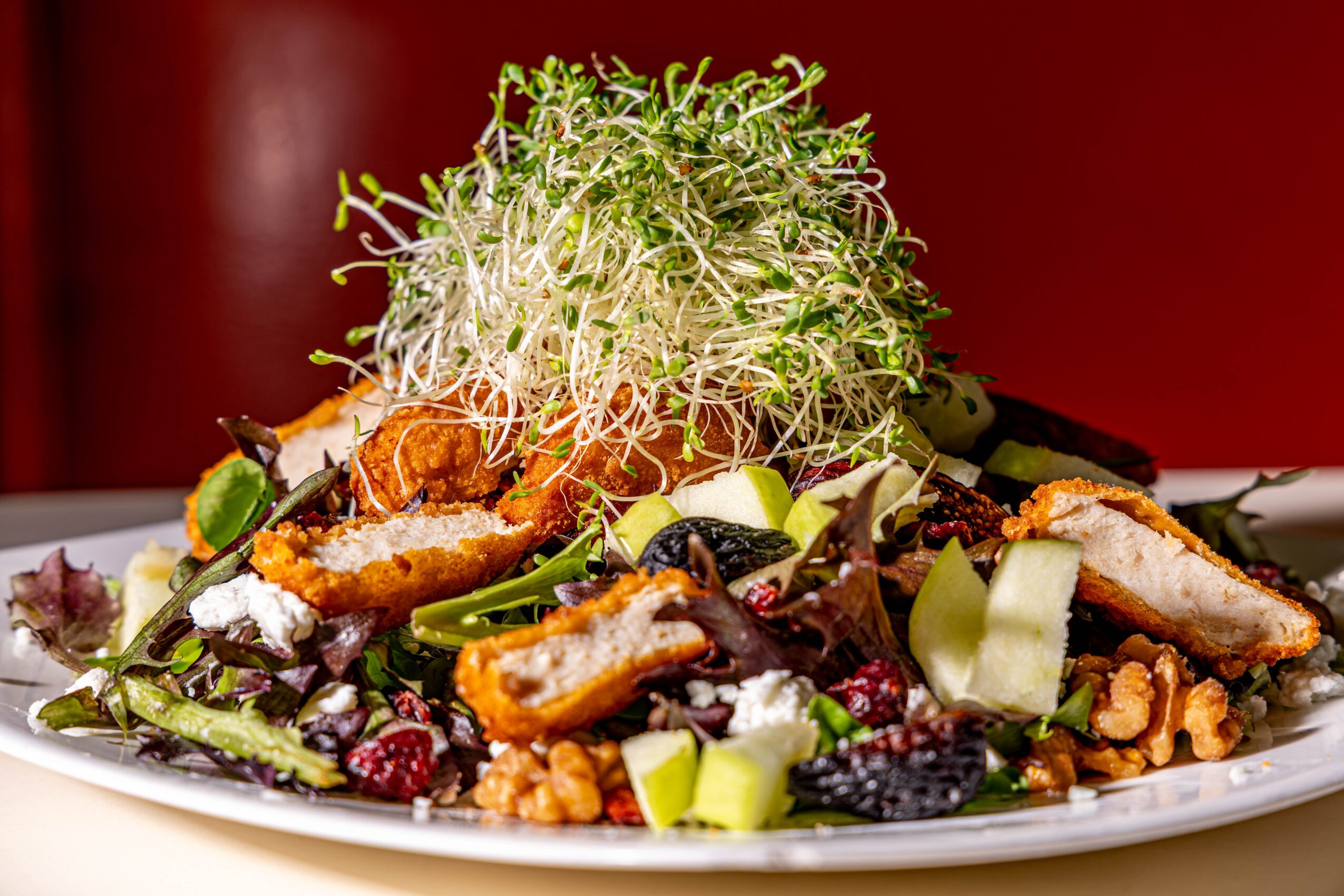 Salad with greens, grilled chicken, nuts, cheese, and vegetables on a white plate against a red background.
