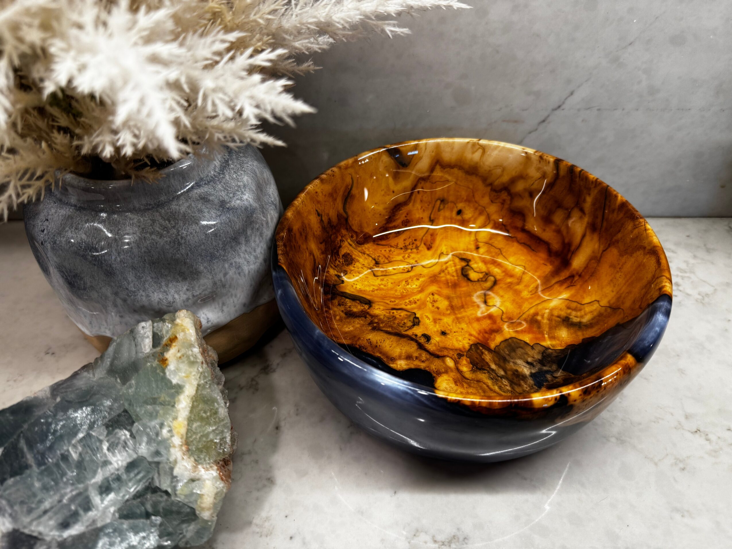 A bowl with a glossy, marbled brown and black interior, placed on a gray surface next to a gray vase with white dried flowers and a large greenish rock.